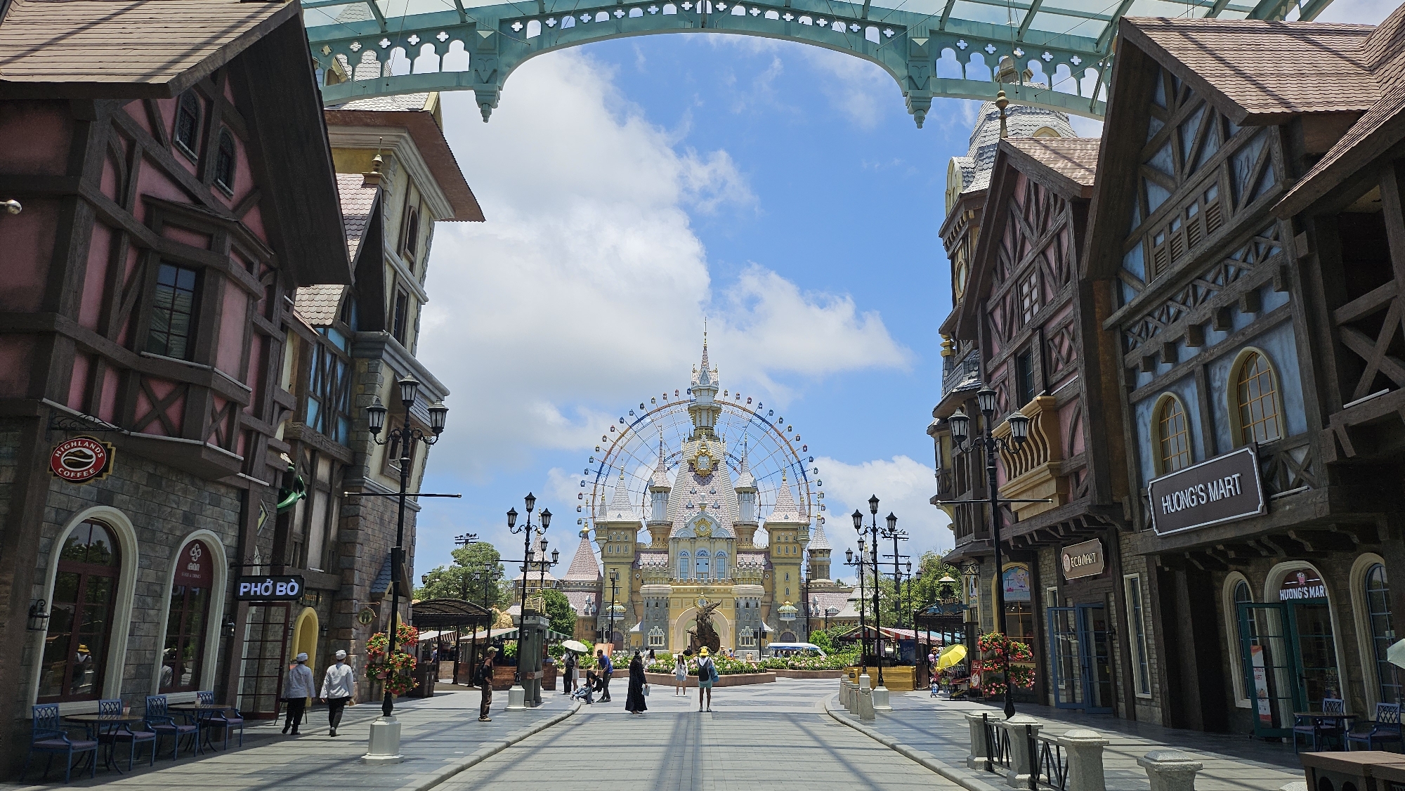 The image depicts a street scene in Sun World, Phú Quốc, Vietnam, designed to resemble a European village. On the left and right sides of the street are multi-story buildings with half-timbered architectural styles, featuring peaked roofs and decorative wooden beams. Signs indicate businesses such as "Highlands Coffee" and "Pho Bo" on the left, and "Huong's Mart" on the right. Paved sidewalks line the buildings, with tables and chairs set up outside some establishments. A lamppost with multiple lights stands on the left sidewalk.
In the distance, at the end of the street, a large Ferris wheel is prominently featured, situated behind a fanciful castle-like structure with multiple spires and towers. The sky above is blue with scattered white clouds. Several people are visible walking along the street and sidewalks, some appearing to be visitors and others possibly staff. The overall atmosphere suggests a themed amusement park or entertainment district.