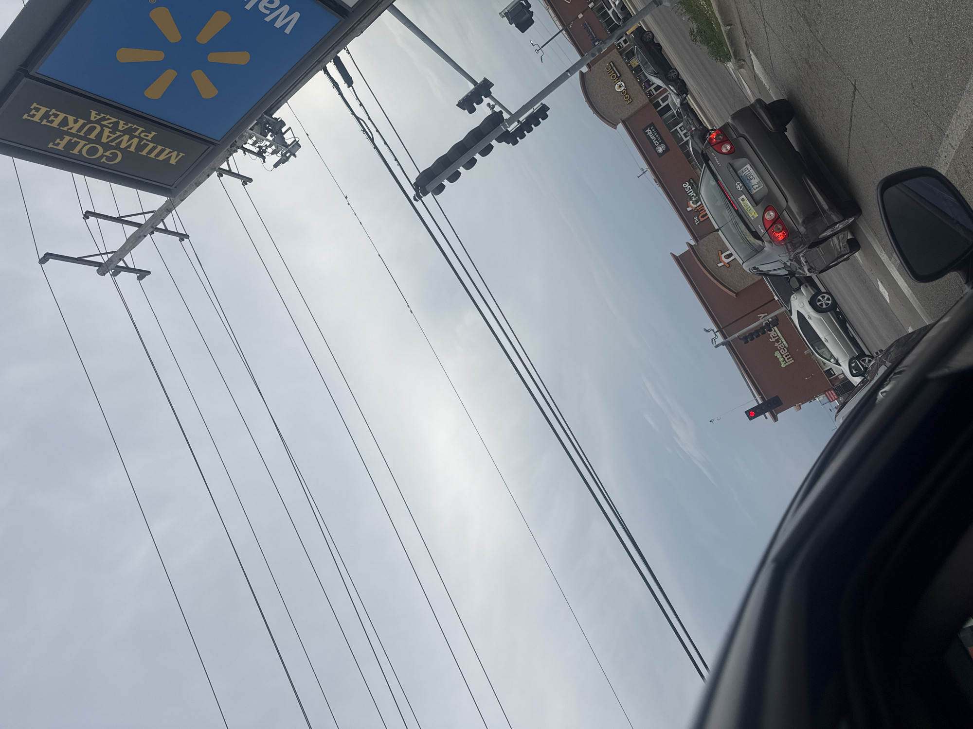 The image is taken from a vehicle at an intersection in Niles, United States of America, looking up and to the right. Overhead, a network of power lines crisscrosses the sky. To the right, a tall signpost displays "GOLF MILWAUKEE PLAZA" in gold lettering on a black background, next to a large blue sign with a yellow Walmart logo. In the distance, traffic lights are visible, one displaying a red light. Several cars are on the road, with a gray sedan in the foreground. Buildings are lined up along the street, including a storefront labeled "EGG N' HOLIC" and another with "Crumbl Cookies". The sky is overcast.