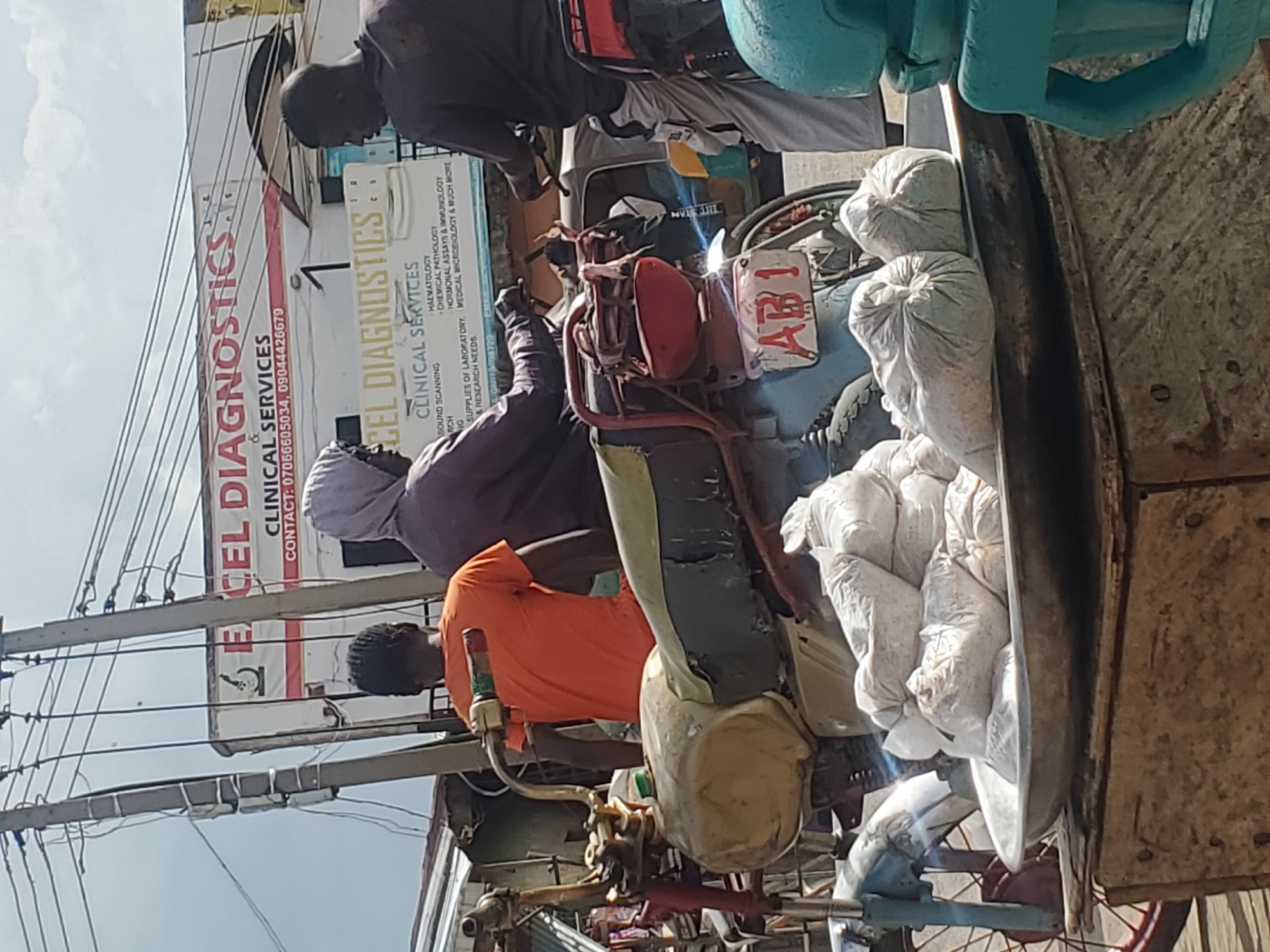 This media file depicts a street scene in Gwong, Nigeria, likely during daytime given the bright sky and lighting. The central focus is a motorized tricycle, commonly known as a Keke Napep, laden with white sacks of goods. Three men are visible around the tricycle. One man, wearing an orange shirt, is standing and appears to be attending to the tricycle's controls or cargo. Another man, in a dark purple top and white headscarf, is seated on the tricycle, looking down. A third man, in dark clothing, is partially visible in the background, also near the tricycle.
In the background, a prominent sign for "EXCEL DIAGNOSTICS & CLINICAL SERVICES" is clearly visible. The sign lists various services offered, including "HAEMATOLOGY," "CHEMICAL PATHOLOGY," "HORMONAL ASSAYS & IMMUNOLOGY," and "MEDICAL MICROBIOLOGY." Contact phone numbers are also displayed. The presence of the diagnostic center and the Keke Napep suggests an urban or semi-urban commercial area.
The Keke Napep itself has a license plate reading "AB1." The sacks appear to be filled with granular or powdery material, possibly agricultural produce or construction supplies. The overall impression is one of daily commerce and transportation in a Nigerian setting, with the men engaged in loading, unloading, or preparing the vehicle for transit. The sky is bright, indicating clear weather.