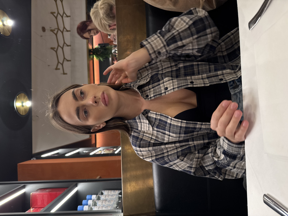 This image depicts a woman seated at a table within an indoor restaurant or bar, identified as being in Verona, Italy. The primary subject, a woman with long brown hair, is wearing a blue, white, and tan plaid button-up shirt, unbuttoned to reveal a black low-cut top, and light blue denim trousers. Her left hand is raised, with fingers touching her neck/ear area, and her gaze is directed towards the upper right portion of the frame with a neutral expression. She occupies a black booth-style seat. The table directly in front of her is covered with a white tablecloth, with a metal utensil visible on the left.
The background features a dark wall with a vertically illuminated shelf system on the left, displaying red items and multiple bottles of San Pellegrino mineral water. A brass-toned partition separates the foreground from a deeper section of the establishment. In this background section, two individuals are partially visible: one woman with red hair, seen in profile, looking downwards, and another person with blonde hair partially obscured behind the partition. Overhead, several circular brass light fixtures with exposed bulbs and a decorative, open-frame hexagonal light fixture are present, with reflections on a perceived reflective ceiling surface. The overall lighting is dim, utilizing accent illumination.