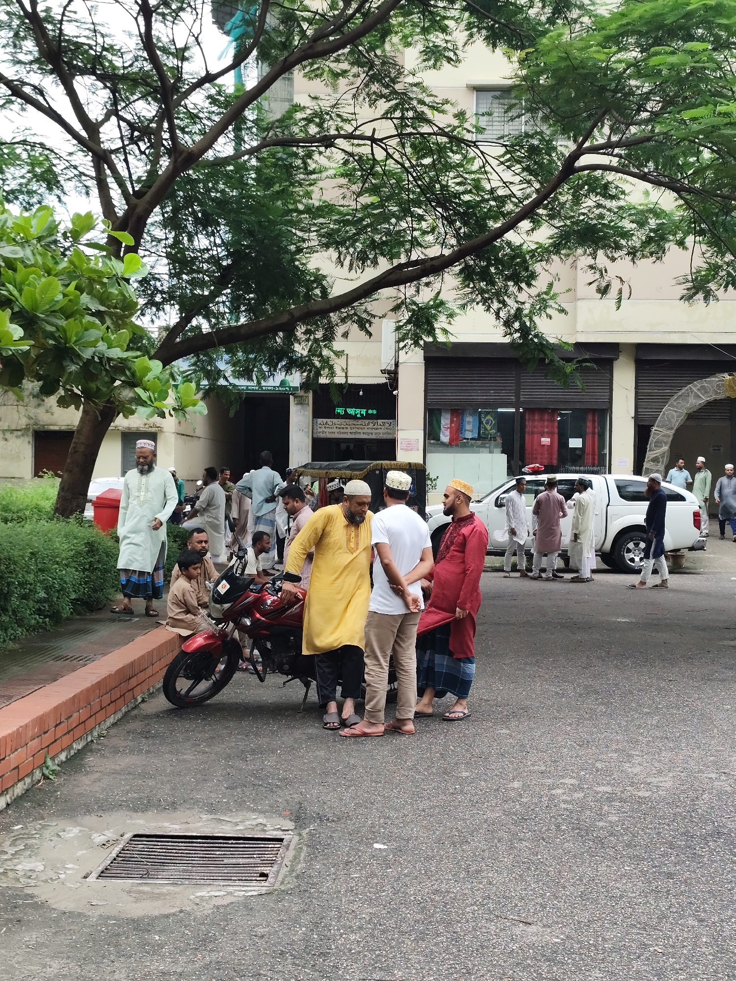 A group of Muslim men in traditional attire are gathered on a paved area in front of a building in Dhaka, Bangladesh. The men, varying in age, are dressed in kurtas, lungis, and kufis, with some wearing colorful caps. The atmosphere appears relaxed, with men standing, talking, and observing their surroundings. A red motorcycle is parked on the side, and a white pickup truck is visible in the background. The environment suggests a public space, possibly near a mosque or a community center, given the attire of the individuals. Greenery from trees partially obscures the building, suggesting an urban setting with some vegetation.