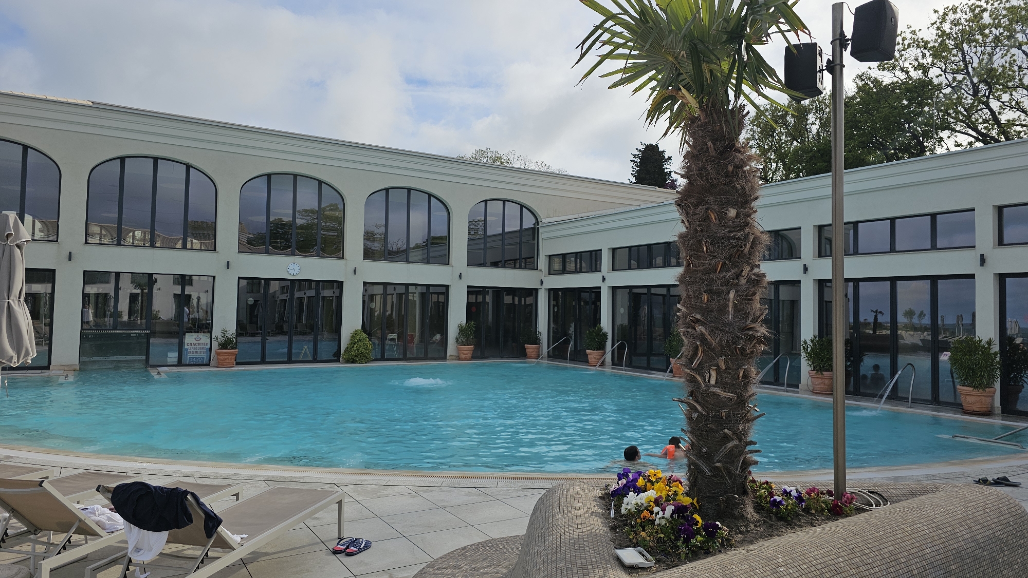 The image shows an outdoor swimming pool area at a building in the city of Varna, Bulgaria. The pool is surrounded by a light-colored building with large arched windows and several potted plants. There are two people swimming in the pool. A palm tree stands near the edge of the pool, adjacent to a flowerbed. Lounge chairs are visible in the foreground, with clothing items on one. A speaker is mounted on a pole near the palm tree. The sky is overcast.