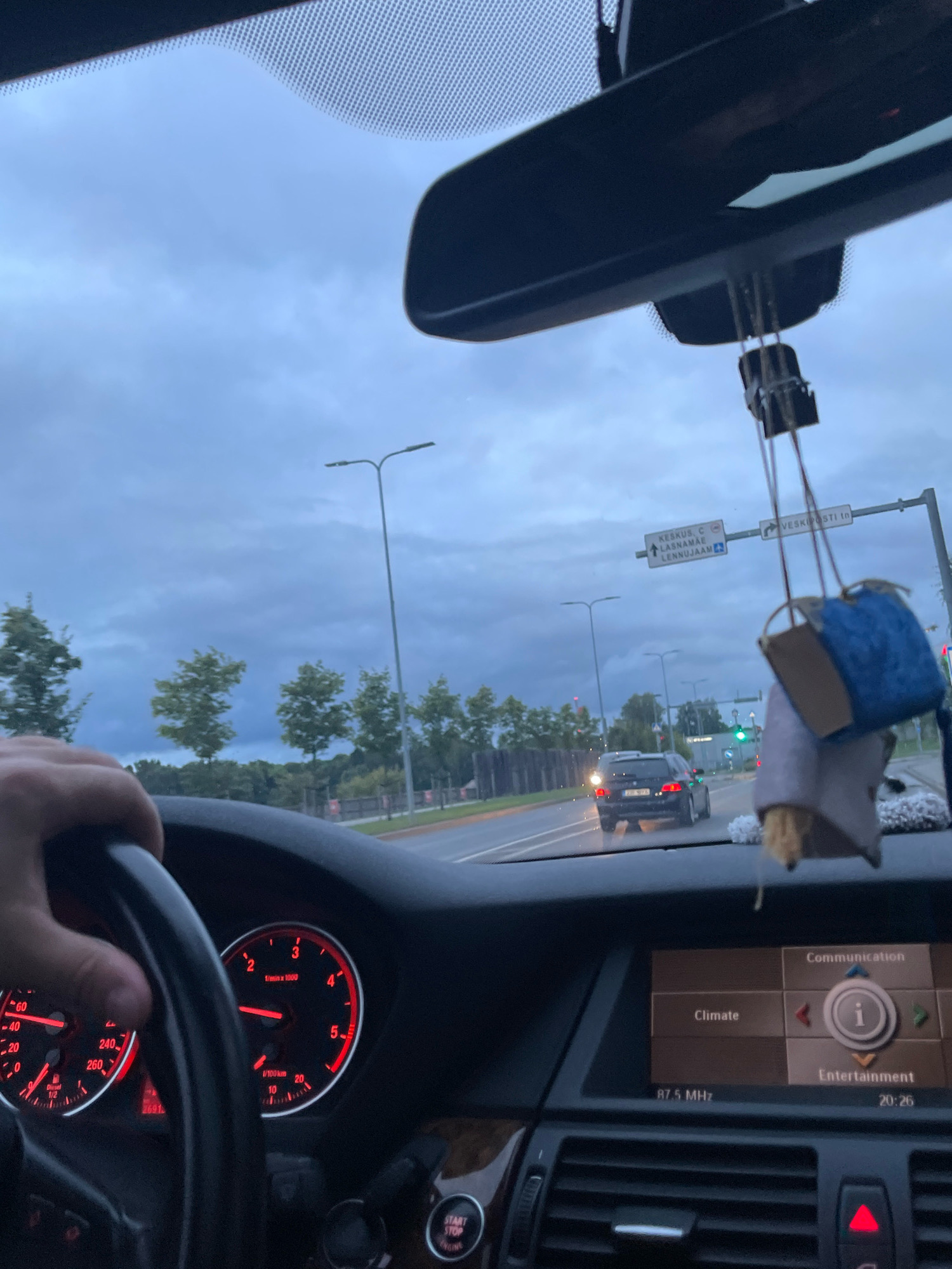 This is a first-person perspective, interior shot of a car driving in Tallinn, Estonia, likely during overcast weather. The driver's hand is visible on the steering wheel. The car's dashboard displays speed and RPM gauges with red illuminated numbers. The central infotainment screen shows "Communication," "Climate," and "Entertainment" options. A GPS display indicates a location in Tallinn with signs for "KESKUS C," "LASNAMÄE," and "LENNUJAAM," which translates to "CENTER C," "LASNAMÄE," and "AIRPORT." The road ahead shows other vehicles, including a black BMW sedan. Traffic lights are visible. A decorative air freshener hangs from the rearview mirror.