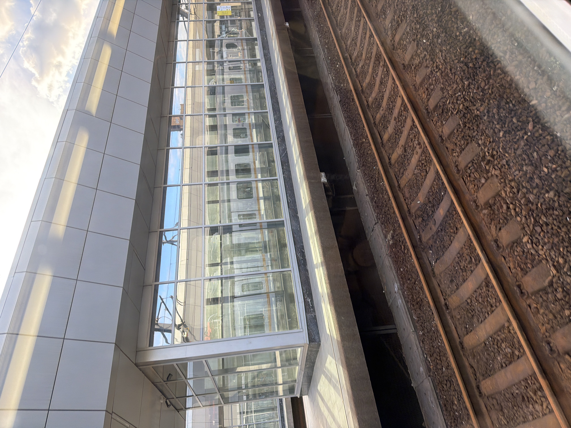 The image depicts a modern building with a glass facade and train tracks. The building is primarily white and gray, with geometric paneling. The glass facade reflects the sky, clouds, and a train. The train appears to be a light color, possibly silver or white, with windows. The train tracks are made of gravel and steel rails, and they are positioned next to the building. The scene is shot from a high angle, looking down at the train tracks and up at the building.
The time of day appears to be daytime, with sunlight casting shadows on the building. The sky is partly cloudy, with some bright white clouds and blue sky visible. The weather appears to be fair. There are no people visible in the image.
Notable details include the reflections in the glass facade, which show the train and the sky. The geometric design of the building is also a prominent feature. The contrast between the modern architecture of the building and the industrial nature of the train tracks creates an interesting visual dynamic.
There is some text visible on the train in the reflection, but it is too blurry to read. There is also some text on a sign in the reflection of the building, but it is also illegible.
The image captures a moment of stillness, with the train appearing to be stationary or moving slowly. The overall mood of the image is one of modern urban life and transportation.