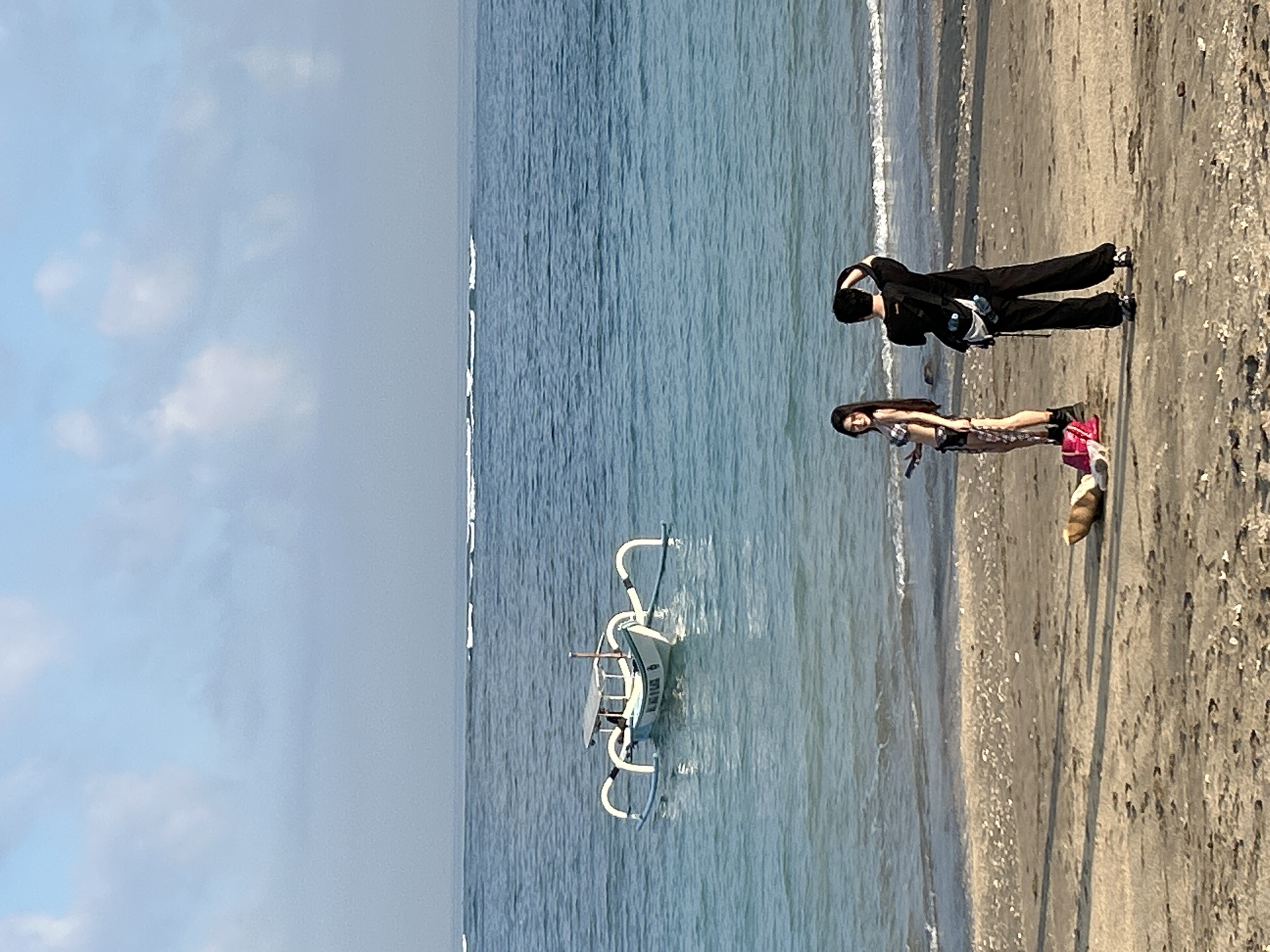 A medium shot captures a sandy beach in Kuta, Indonesia, with two individuals near the waterline. A woman in a black and white checkered bikini stands facing left, holding a phone, with a pink bag and what appears to be a light brown bag with a strap on the sand near her feet. To her right, a person in a black t-shirt and black pants faces away from the camera, with a crossbody bag, and is holding their right hand to the back of their head, possibly adjusting their hair or taking a picture. In the background, the tranquil ocean stretches to the horizon, with faint white waves breaking further out. A traditional Balinese fishing boat, white with blue outriggers, floats on the calm blue water. The sky is a pale blue with scattered white clouds. The sand on the beach is a light brown, interspersed with small white shells and footprints. The lighting suggests it is daytime, likely morning or late afternoon, with soft shadows cast on the sand.