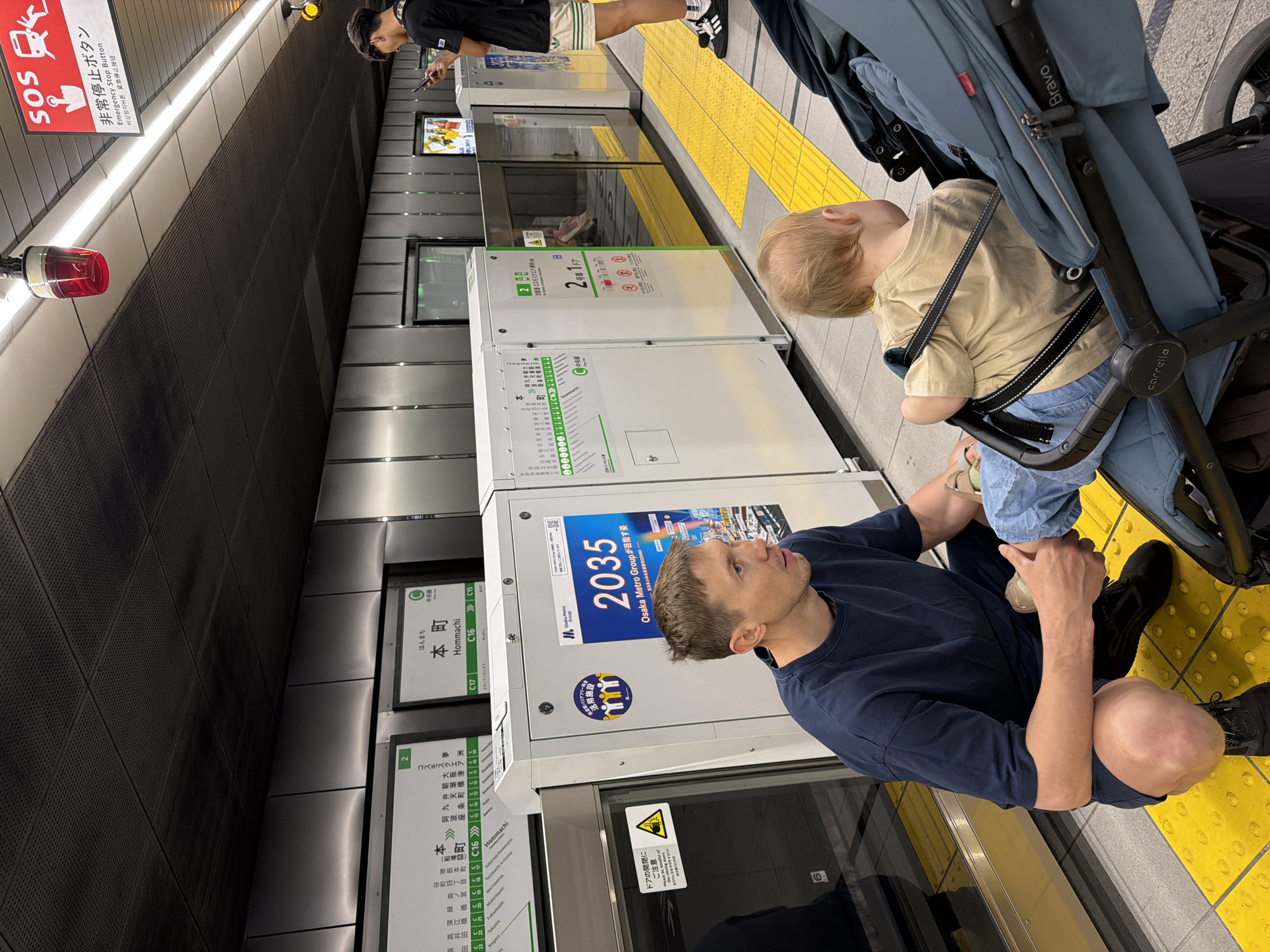 The image captures a scene on a subway platform, likely in Japan, given the visible text. In the foreground, a man in a dark blue t-shirt and shorts kneels on the yellow tactile paving of the platform. He is positioned next to a blue baby stroller, in which a young child with blonde hair, wearing a light beige t-shirt and jeans, is seated and strapped in. The child appears to be looking towards the man or down. The stroller is a "Carrello Bravo" model.
Further down the platform, another individual, partially visible, stands with their back to the wall, engaged with a smartphone. The platform features modern platform screen doors, behind which train tracks can be partially seen. On one of the screen doors, a large blue sign displays "2035 Osaka Metro Groupが目指す姿" (Osaka Metro Group's vision for 2035), along with a sticker indicating "バリアフリー活用施設" (Barrier-free facility). Another sign prominently displays "本町 Hommachi C16", identifying the station as Hommachi on the Chuo Line (中央線 C). A subway map on a door lists several station names, including "谷町四丁目", "堺筋本町", "森ノ宮", "深江橋", and others.
An emergency stop button, labeled "SOS 非常停止ボタン Emergency Stop Button" in multiple languages (Japanese, English, Korean, Chinese), is mounted on the wall with a red light above it. Another sign on a door warns, "ドアの開閉にご注意 Please be mindful of the doors opening/closing." The platform is brightly lit, suggesting an indoor setting. The atmosphere appears calm as people wait for a train.