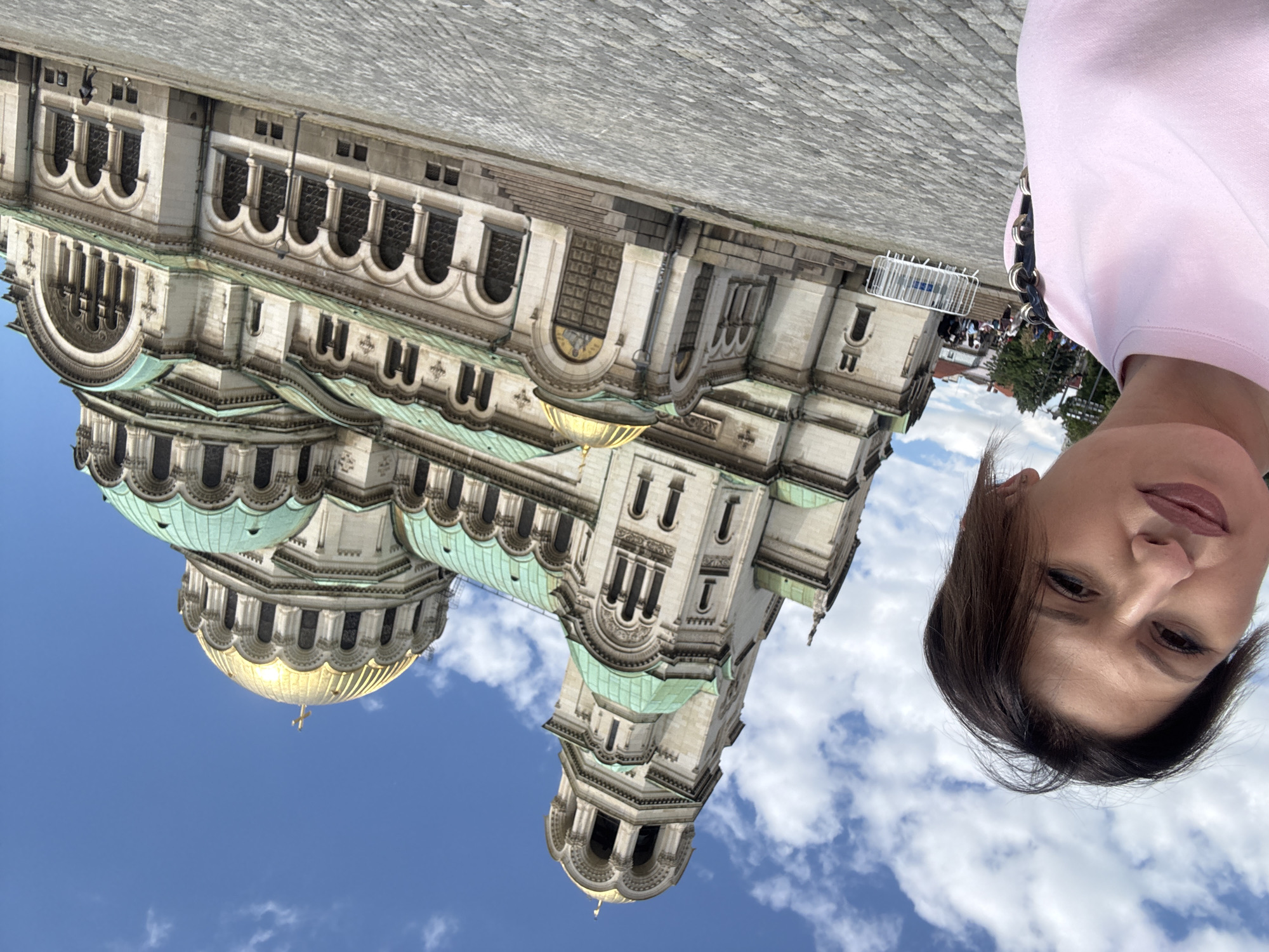 A bright outdoor vertical shot captures a woman taking a selfie in front of a grand architectural landmark under a clear blue sky. The image is oriented with the woman's head on the right side, looking directly at the camera, while a large, ornate building dominates the left and center.
The woman, with dark hair parted to the side, features a neutral expression with a slight pout and visible eye makeup. She is wearing a pink top, the shoulder of which is visible on the right, and a dark handbag with a metallic chain strap rests on her shoulder.
The imposing building, identifiable as the Alexander Nevsky Cathedral in Sofia, Bulgaria, features intricate stone architecture with multiple domes. Several large, golden domes shine brightly in the sunlight, contrasting with numerous smaller domes and sections of the roof that are covered in green patina. The cathedral's facade showcases arched windows, decorative stone carvings, and various architectural tiers.
The setting appears to be a public square or cobblestone plaza, with the textured ground visible in the bottom right corner. In the distant background, near the building, there are faint indications of other people and some metal barriers or fencing, suggesting a public area or event. The sky is bright blue with scattered white cumulus clouds, indicating fair weather and daytime conditions, likely midday or afternoon given the strong illumination on the building. No visible text is present in the image.