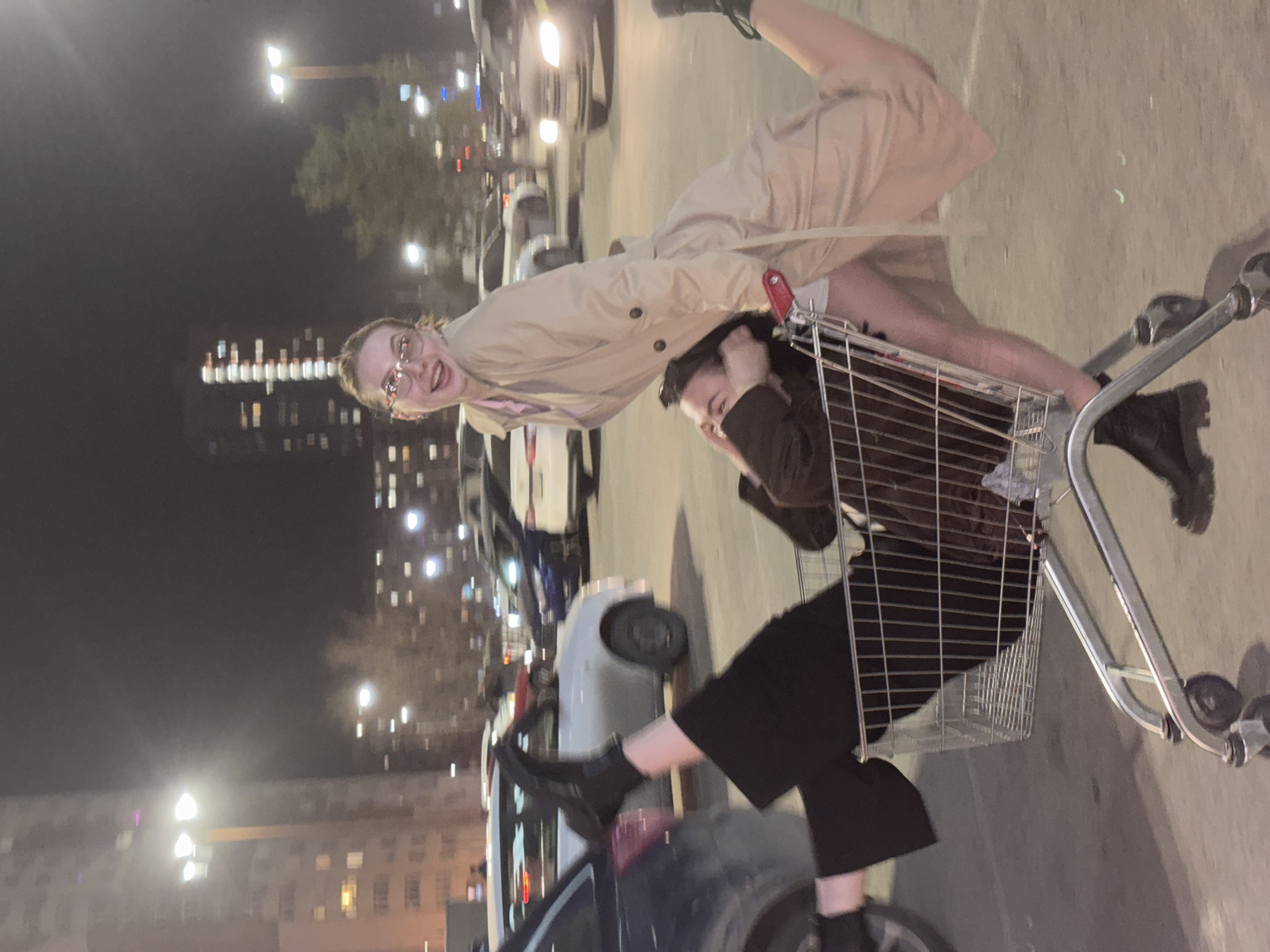 The image depicts a nighttime scene in an urban parking lot in Perm, Russia, featuring two young individuals engaged in a playful activity with a shopping cart.
One person, positioned at the rear of a metal shopping cart, is standing with bent legs and appears to be pushing or riding on it. They wear glasses, a light-colored trench coat, and dark boots, looking directly at the camera with a wide, joyful smile.
The second individual is seated inside the wire basket of the shopping cart, dressed in dark attire, including a jacket, trousers, and boots. Their face is partially obscured, but their posture suggests they are bracing themselves for movement.
The background shows several parked cars and illuminated streetlights, indicating the dark hours. Beyond the parking lot, multi-story buildings with numerous lit windows are visible, confirming an urban setting. The ground surface is dark asphalt or concrete. The overall mood conveyed is one of fun and youthful exuberance. No visible text is present in the image.
