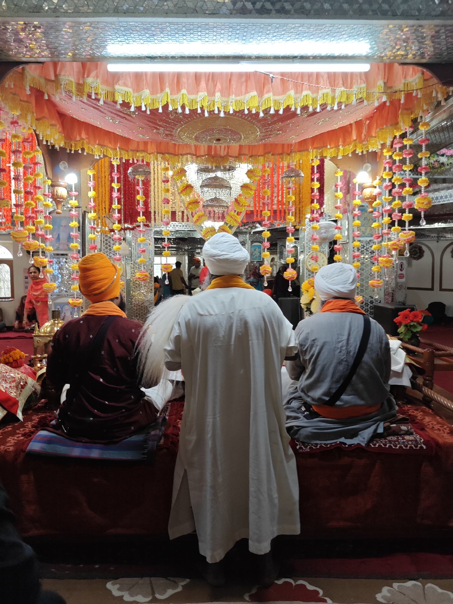 This image depicts the interior of a vibrantly decorated Gurudwara in Kasol, India, bustling with religious activity. In the foreground, three men are seated on a raised, ornate platform, viewed from behind, facing what appears to be an altar or sacred area. The man on the left wears an orange turban and a maroon traditional garment with an orange sash. The central figure is dressed in a white turban and a long white robe, with a chaur sahib (fly-whisk) resting on his shoulder, signifying a role in a religious ceremony. The man on the right wears a white turban, a grey robe, and an orange sash, with a black strap across his back.
The setting is lavishly adorned with numerous hanging garlands of artificial flowers in shades of yellow, pink, and red, creating a festive and sacred ambiance. A prominent peach-colored canopy, embellished with yellow and white tassels, is suspended above the platform, complementing the shimmering, patterned ceiling panels and bright fluorescent lighting. In the background, other devotees are visible, including a woman in an orange-red outfit standing on the left. Decorative elements such as a golden, dome-shaped object on the platform, intricate metal hangings, and vases with red flowers contribute to the rich decor. The platform itself is covered with plush maroon and red patterned fabrics. The individuals present are likely participating in or observing a religious service or prayer. The word "IKON" is partially visible on a piece of fabric on the platform. The artificial lighting suggests the time of day is either evening or that the interior relies on constant illumination.