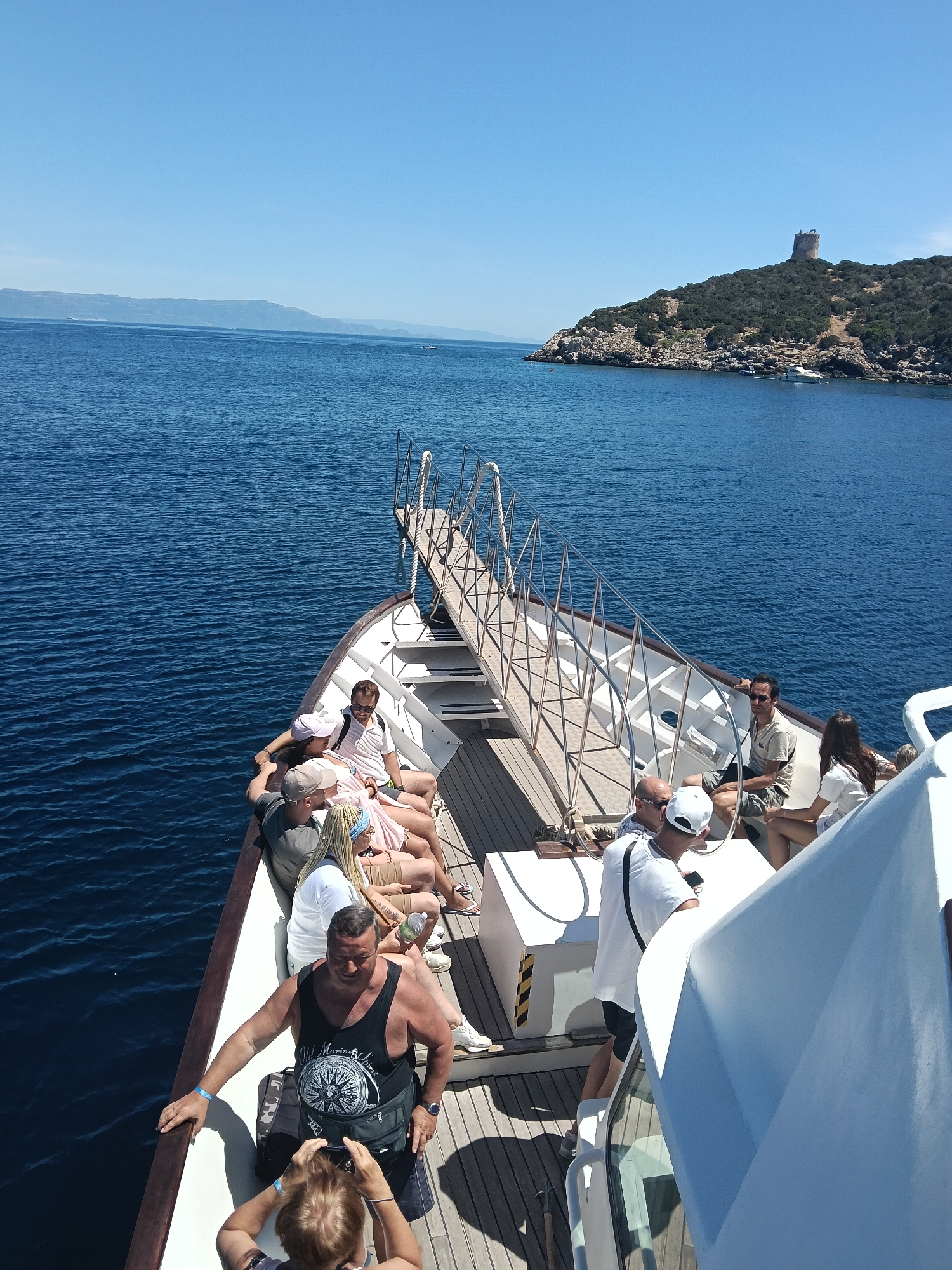 The image shows a group of people on a boat in the sea. The boat has a wooden deck and a metal railing. Some people are sitting on the deck, while others are standing. In the background, there is a coastal landscape with a tower on a hill. The sky is clear and blue, and the water is calm and blue. The scene appears to be a tourist excursion.