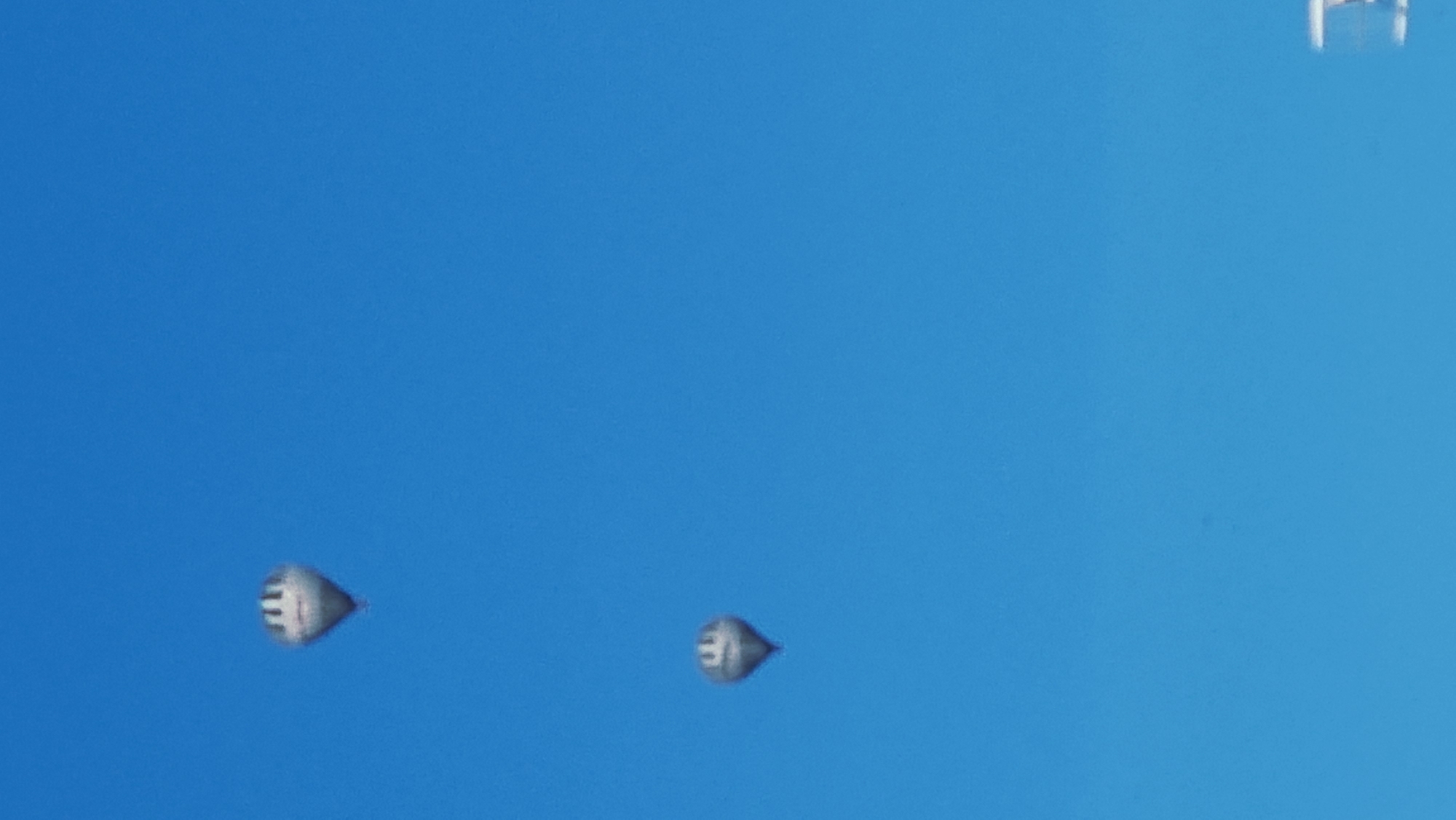 The image shows two hot air balloons against a clear blue sky. The balloon on the left is silver with black stripes and appears to be slightly larger. The balloon on the right is also silver with black stripes and is positioned lower and to the left of the first balloon. The scene is a wide shot, with the sky taking up most of the frame. No other objects or people are visible. The location is not identifiable from the image alone, but the prompt specifies the city of Sant Joan Les Fonts, Spain.