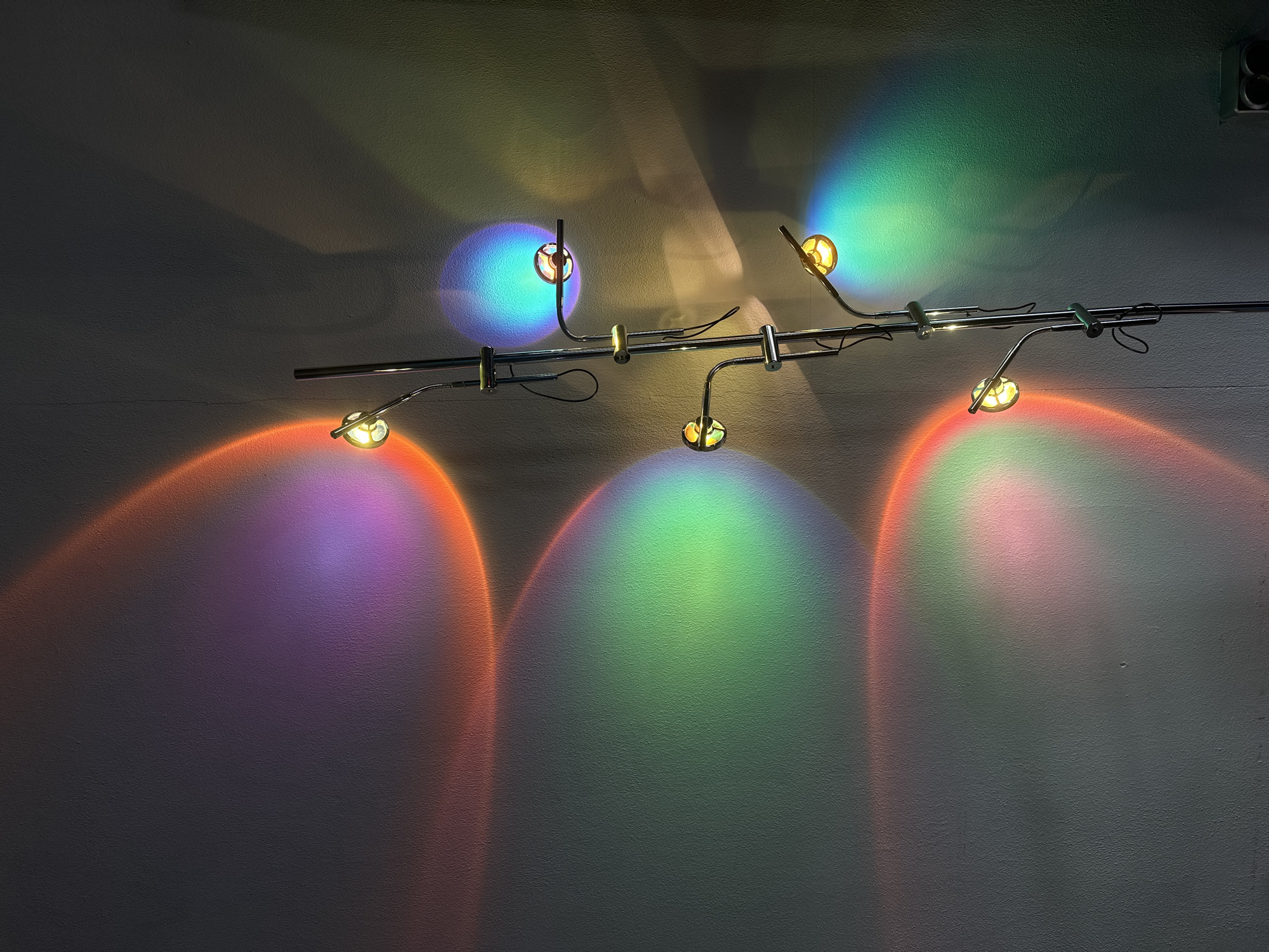 The image depicts a modern track lighting fixture mounted on a textured wall, likely indoors. The fixture consists of a metal rail with several adjustable spotlights. These spotlights are actively casting colorful beams of light onto the wall, creating abstract patterns of red, orange, purple, blue, and green. The lighting effect resembles a sunset or a mood lighting installation.
There are no people visible in the image. The focus is entirely on the lighting fixture and its colorful projections. The wall itself is a neutral, light gray color with a noticeable texture, suggesting it could be a concrete or stucco surface. The shadows cast by the fixture are sharp, indicating a direct light source.
The overall impression is one of a contemporary interior space, possibly a gallery, studio, or a stylishly decorated room. The colorful lights create a vibrant and artistic atmosphere. There is no visible text in the image, and no clues about the time of day or weather conditions, as it's an indoor scene. The location context of Moscow, Russia, does not provide any specific visual cues within this image.