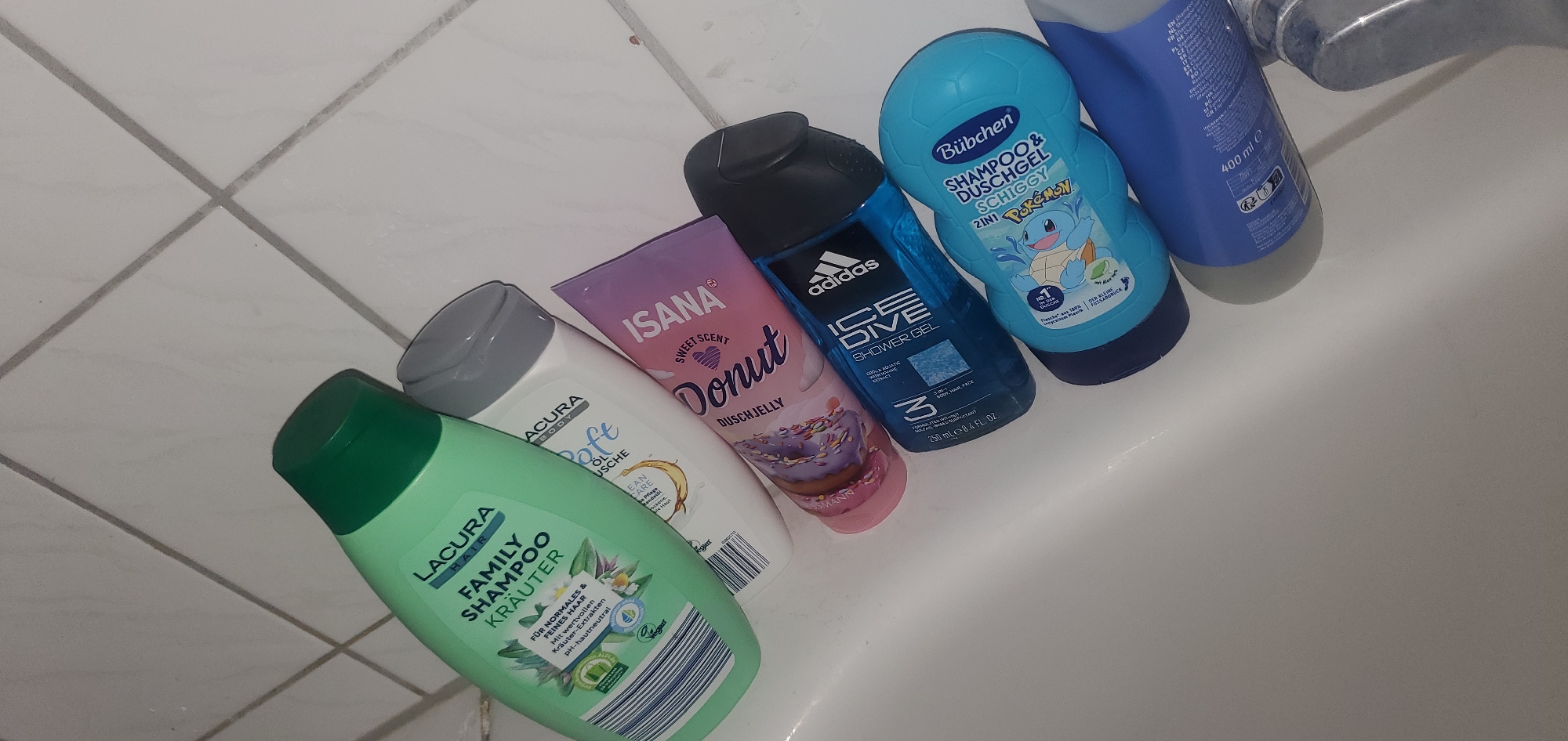 Six bottles of shampoo and shower gel are arranged in a bathtub. The bottles include Lacura Family Shampoo Kräuter, Lacura Body Soft Oil Dusche, Isana Sweet Scent Donut Duschjelly, Adidas Ice Dive Shower Gel, Bübchen Shampoo & Duschgel Schiggy (Pokémon themed), and another blue bottle of shampoo, possibly by Adidas, with "400 ml" visible. The background is a white tiled wall. The image was taken in Sasbach, Germany, in a bathtub.