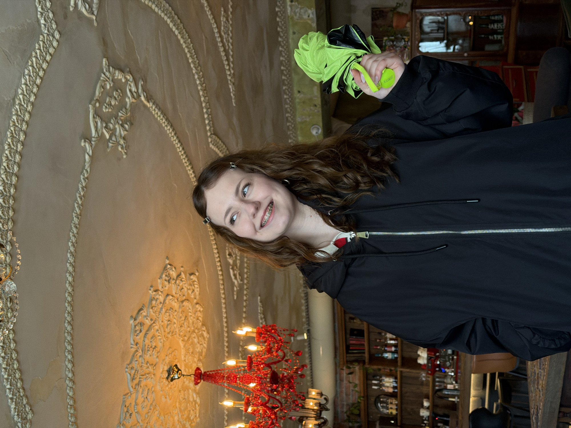 A young woman with braces, wearing a black jacket and holding a folded green and black umbrella, is indoors. The background features an ornate wall with decorative molding and a large red chandelier. Shelves filled with various items, including bottles, are visible behind her. This photograph was taken in the city of Ryazan, Russia.