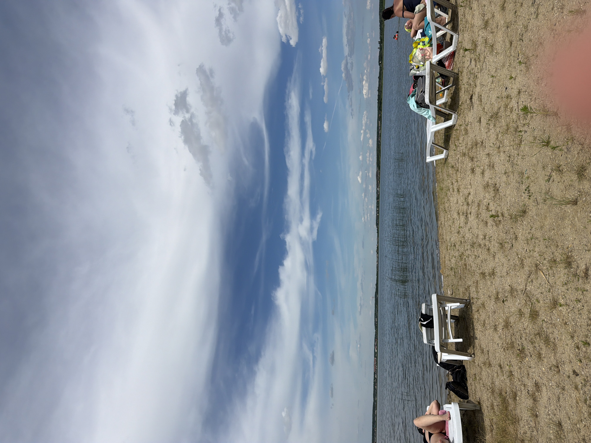 The image depicts a sandy beach area adjacent to a large body of water, likely a lake or reservoir. The sky is partly cloudy with a mix of white and grey formations. In the foreground, several white plastic lounge chairs are set up on the sand. One chair on the left has a person lying on it, wearing dark swimwear. Another chair in the middle has towels and clothing draped over it. To the right, two more lounge chairs are occupied by people. One person is lying down, and another is sitting up, wearing a dark top. The water is calm and reflects the sky. Along the distant shoreline, a line of trees and buildings is visible, suggesting a settlement. Reeds or tall grass grow in the water near the shore. The location is identified as Kremenkul', Russia, indicating a recreational area by a body of water.