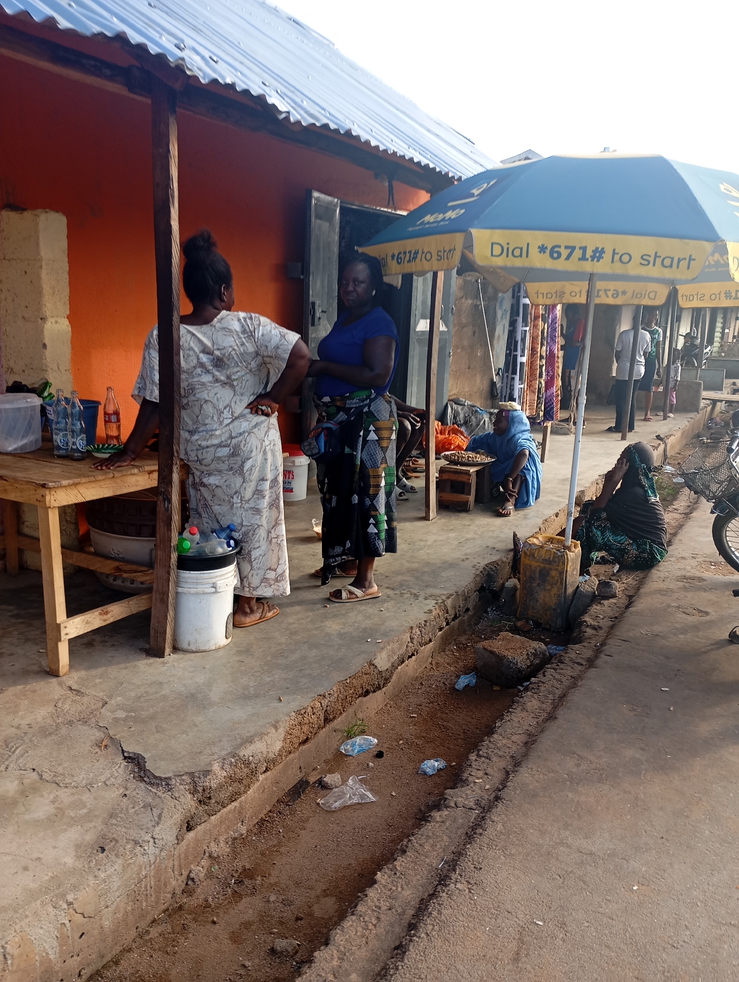 The image depicts an outdoor marketplace scene in Shendam, Nigeria. In the foreground, a concrete ditch runs along the side of a paved walkway, with some debris visible within it. Several people are present in the scene. Two women are standing near a wooden table laden with bottles and containers. To the right, under a blue and yellow umbrella advertising a phone service, another woman is seated on the ground, talking on a cell phone. Further back, several other individuals are visible, including some standing and others seated, engaged in various activities within the market. The backdrop includes a building with an orange-colored wall and a corrugated metal roof, as well as stalls displaying various goods. The overall atmosphere suggests a typical day at a local Nigerian market.