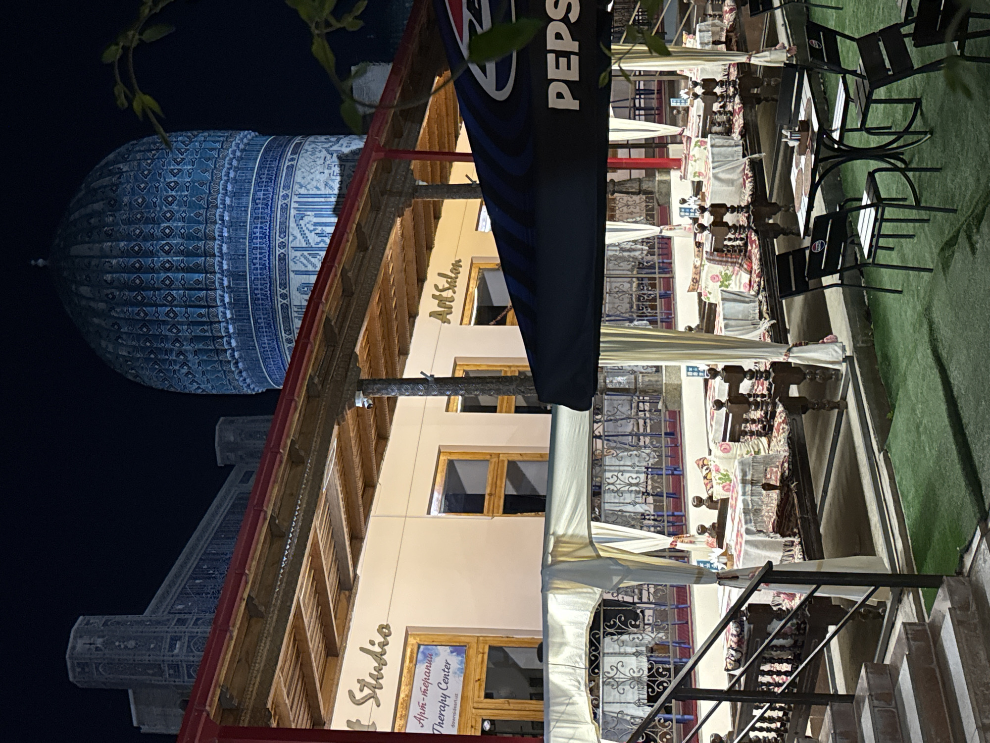 The image depicts a nighttime scene in Samarkand City, Uzbekistan. In the background, the turquoise-domed Gur-e-Amir mausoleum is visible. In the foreground is a building with "Studio Art Salon" signage. Tables and chairs are arranged as if for outdoor dining, with some tables set with tablecloths. A Pepsi banner is partially visible, and vegetation is seen at the top of the frame. Stairs lead up to the restaurant area. The overall lighting suggests it is evening.