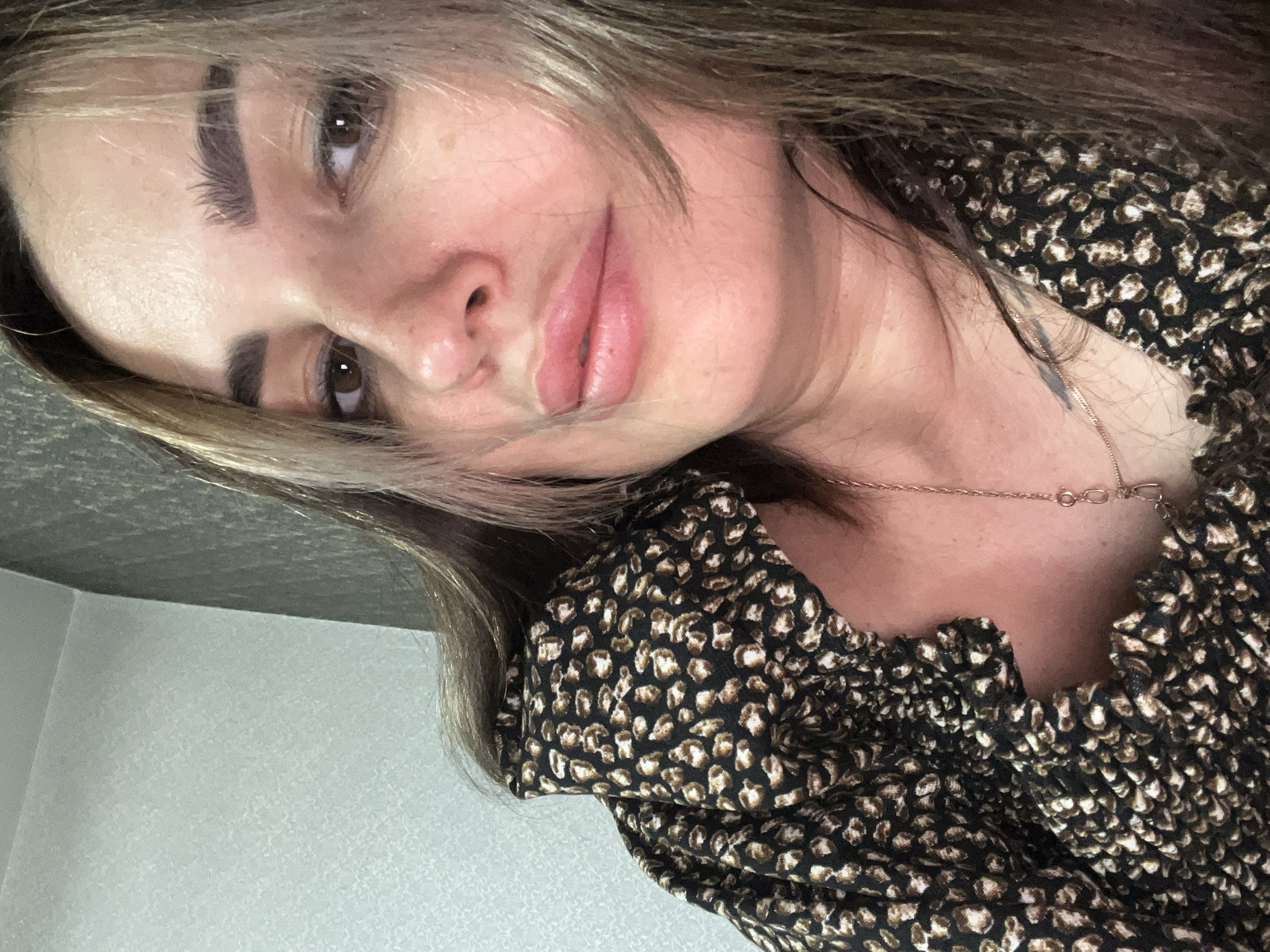 A close-up, head-and-shoulders selfie shows a woman with long, wavy brown hair with lighter streaks. She has dark brown eyes, thick eyebrows, and full lips that appear to be subtly glossed. Her skin has a warm, fair tone with a few faint freckles visible on her cheek. She is wearing a black top with a small, repeating brown and white animal print pattern, and a delicate gold necklace with a small pendant. The background consists of a textured, light-colored wall on the left and a darker, patterned wall on the right, suggesting an indoor setting. The lighting is soft, casting gentle shadows and highlighting her features. The overall impression is that of a candid, casual portrait.