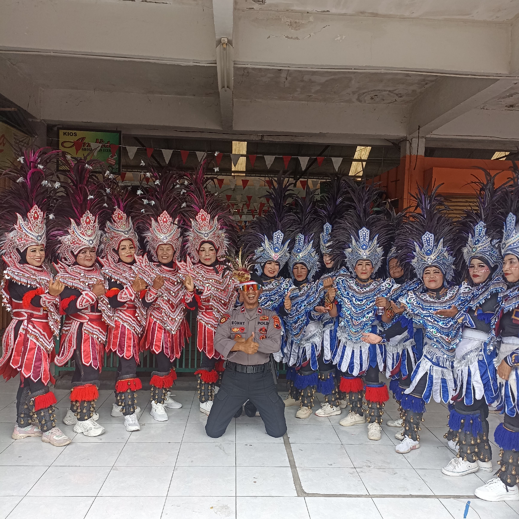 The image depicts a group of individuals dressed in elaborate, colorful costumes, likely for a performance or parade, standing in front of a building with an overhang. The costumes feature large feathered headdresses, ornate jackets with shimmering details, and skirts adorned with bells and fringe. There are two distinct color schemes for the costumes: red and silver, and blue and silver.
In the center of the group, a male individual in a police uniform is kneeling and giving a thumbs-up. He is wearing sunglasses and a band on his head. The police uniform has the name "RONNY" and the word "POLRI" visible on the chest.
The background shows a building with a tiled floor. A sign with the word "KIOS" is visible on the left side of the building. Several triangular flags, alternating red and white, are strung across the top of the background. Plants with dark red and purple foliage are positioned in front of the building. The scene appears to be outdoors, possibly in an urban environment. The presence of "POLRI" on the uniform indicates the location is in Indonesia. Based on the provided context, this scene is located in the city of Magelang, Indonesia.