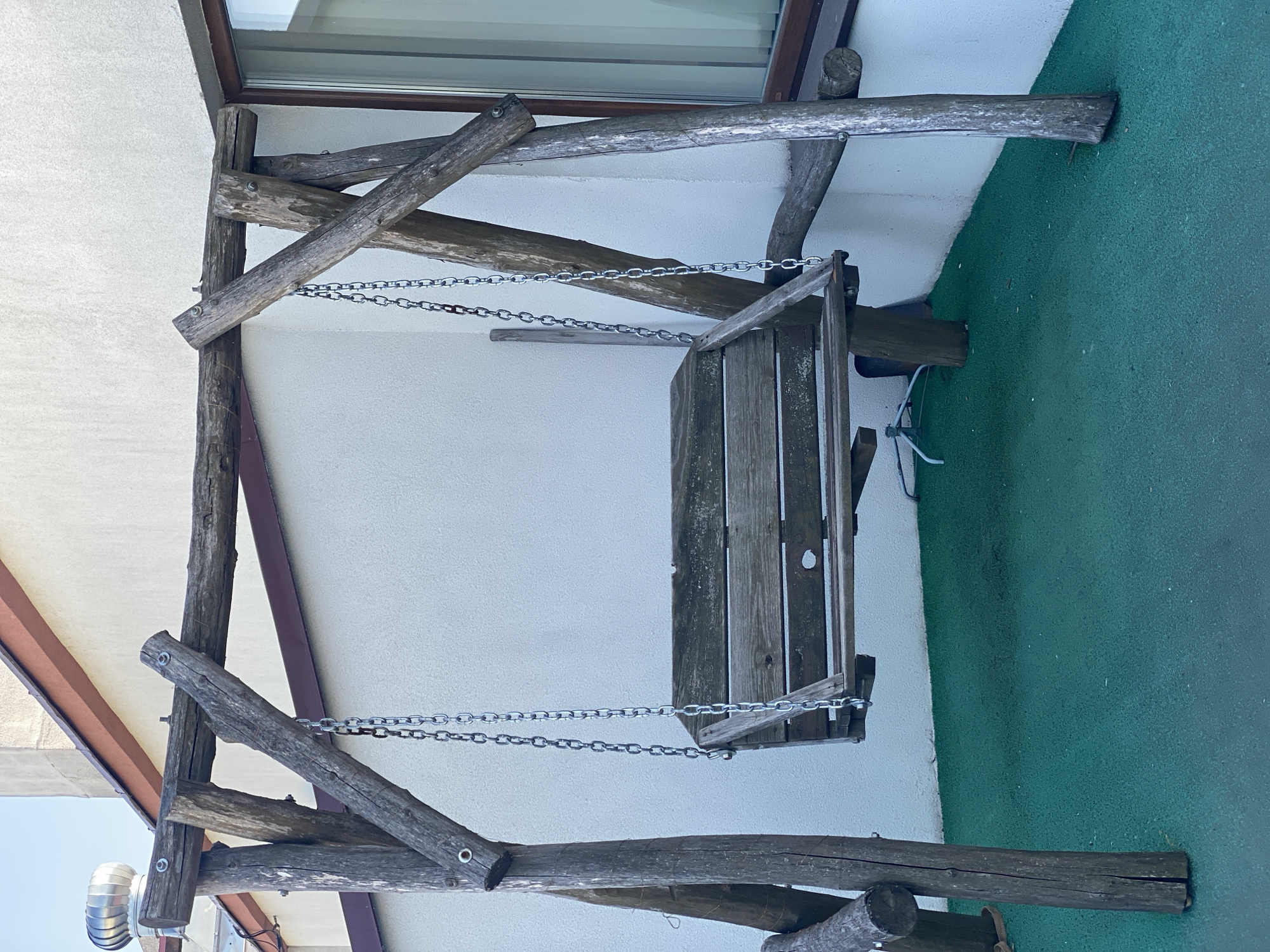 A rustic wooden swing set is positioned on a green, textured surface, possibly artificial turf, adjacent to a white building with a reddish-brown trim under a clear blue sky. The swing set is constructed from rough-hewn logs, with the main horizontal support beam connected to two diagonal A-frame legs on each side. A wooden swing seat, made of planks, is suspended by two metal chains from the horizontal beam. The building features a window with dark framing and blinds. The overall scene appears to be an outdoor area, likely a courtyard or patio. The photograph was taken in Chisinau, Moldova.