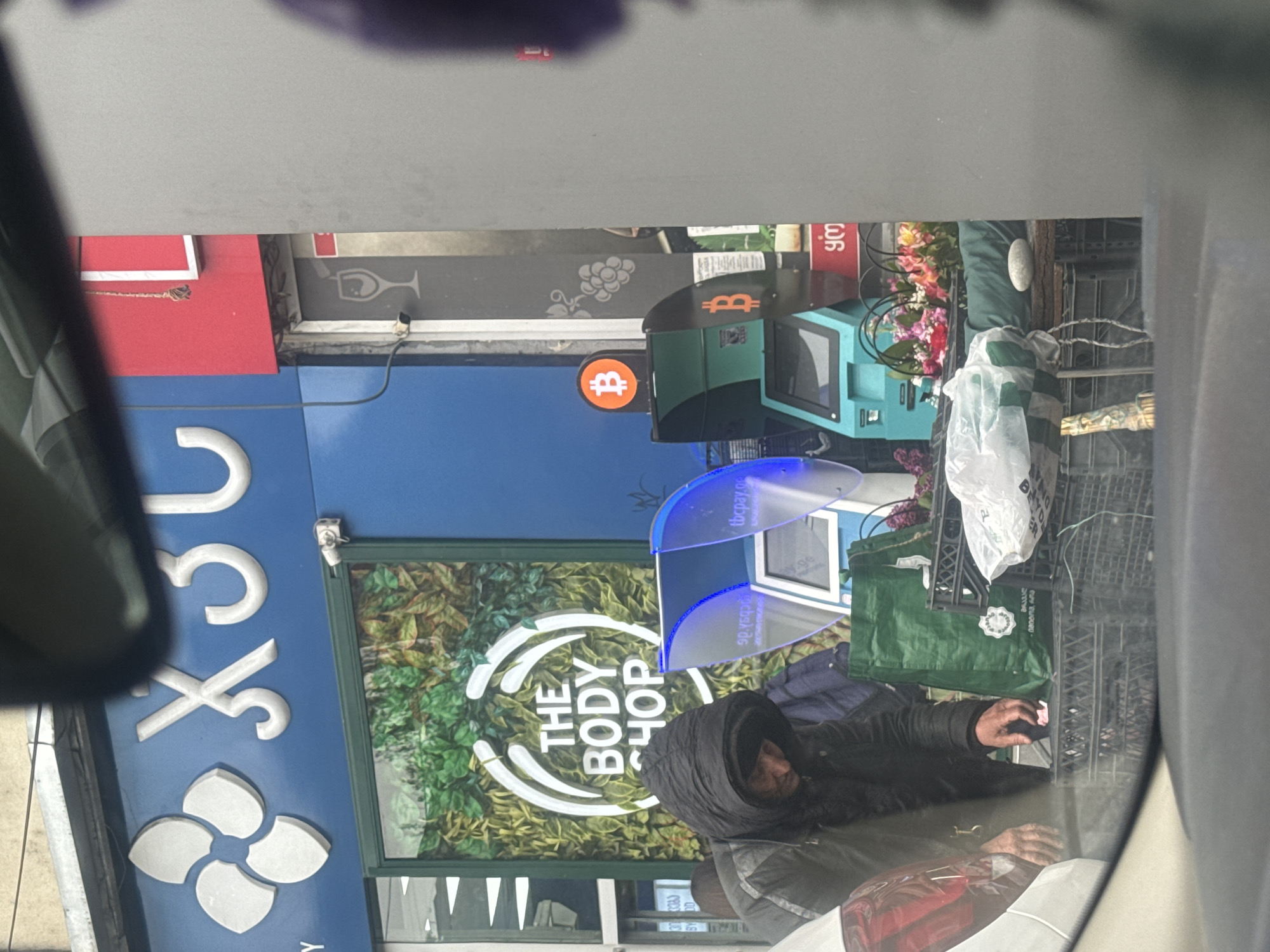 The scene, captured from inside a car, shows the exterior of a Body Shop storefront located in Tbilisi, Georgia. The storefront features the Body Shop logo and signage, with Georgian script visible on signage in the background and on the ATM. A Bitcoin ATM is visible next to the Body Shop. A person wearing a hooded jacket is standing in front of the Body Shop. Near the pedestrian there are flowers, crates, and bags. The picture is taken through the car's windshield.