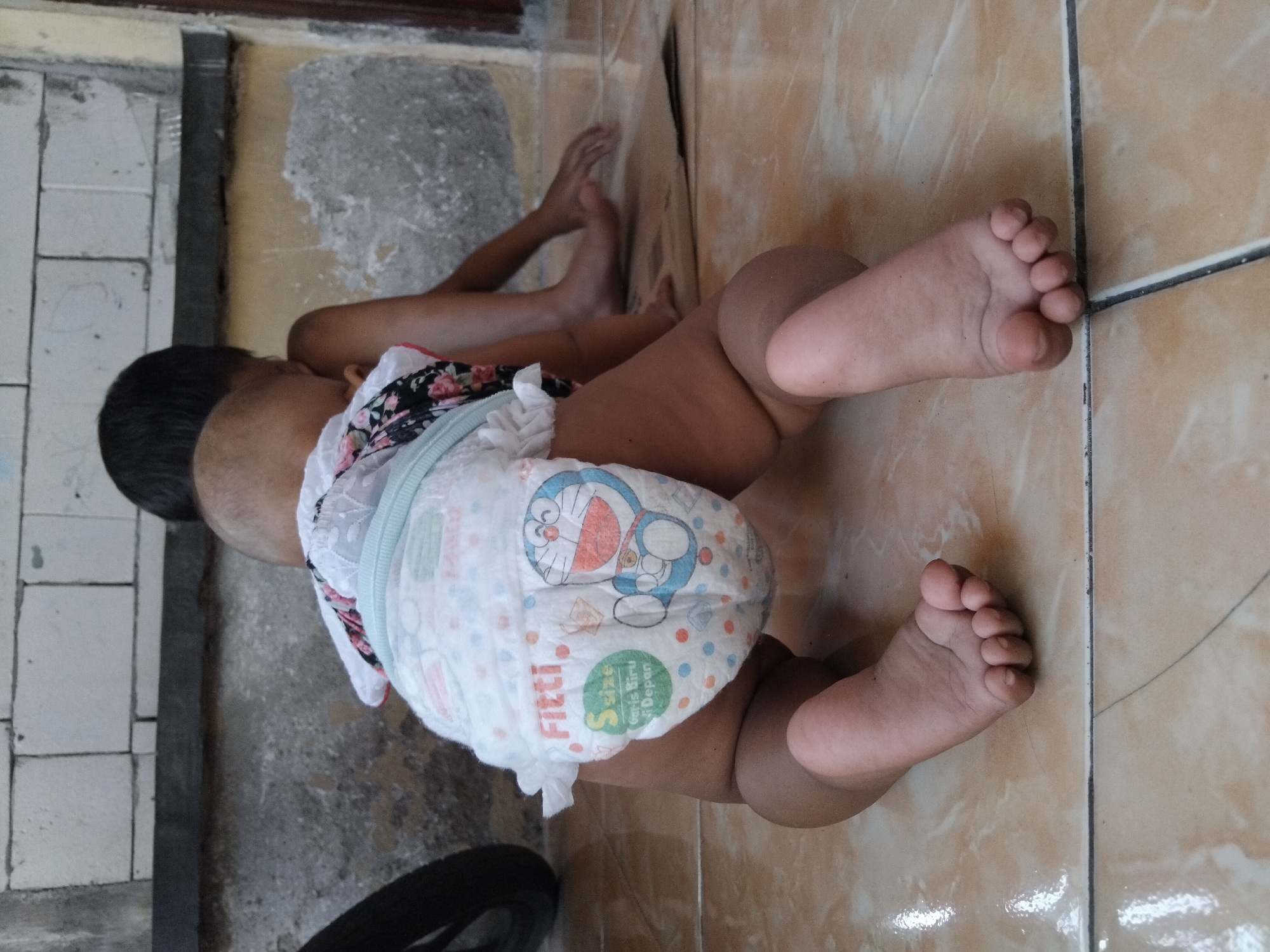 A baby is crawling on a tiled floor. The baby is wearing a diaper with a Doraemon print. The baby's legs and feet are visible. The scene appears to be indoors, with some exposed brick and concrete in the background. The floor tiles are a light brown color. There is no indication of the time of day or weather conditions. The location is identified as Kepuh Kiriman, Indonesia. The diaper has visible text that says "Fitti" and "S size".
