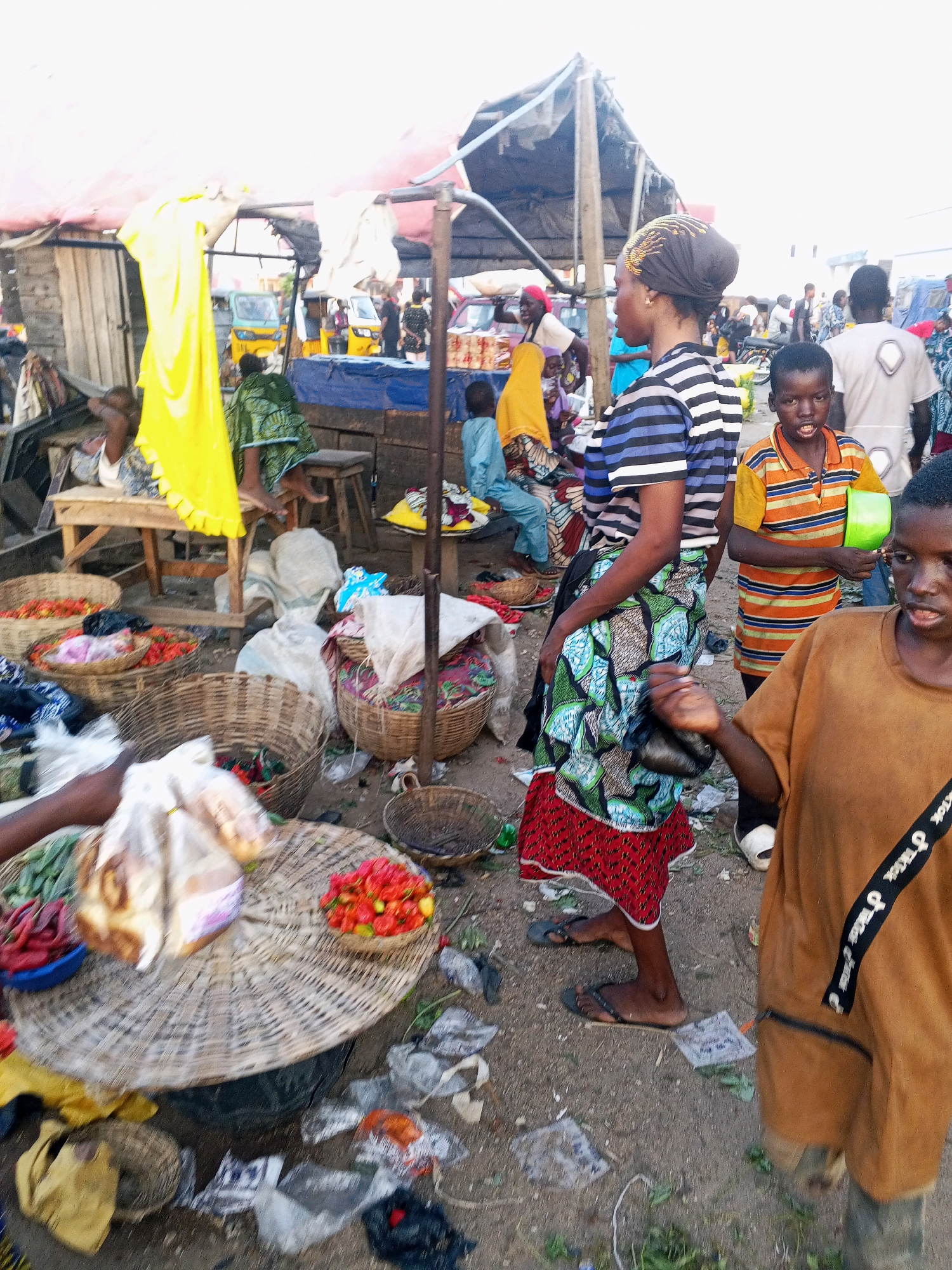 The image depicts an outdoor market scene in Lokoja, Nigeria, characterized by a lively, somewhat chaotic atmosphere. Several individuals, including women, men, and children, are present. A woman in the foreground wears a headscarf and a horizontally striped t-shirt paired with a vibrantly patterned skirt and flip-flops. To her right, a young boy in an orange and yellow striped shirt holds a green bowl. Another young boy in a tan t-shirt stands to the far right, his face partially obscured.
In the background, various stalls are set up under makeshift coverings, with goods like produce, baskets of food, and fabrics displayed. Some individuals are seen tending to their wares or interacting with others. The ground is a mixture of dirt and scattered debris, suggesting a busy and perhaps unkempt environment. The overall impression is a snapshot of daily commerce in a Nigerian market.