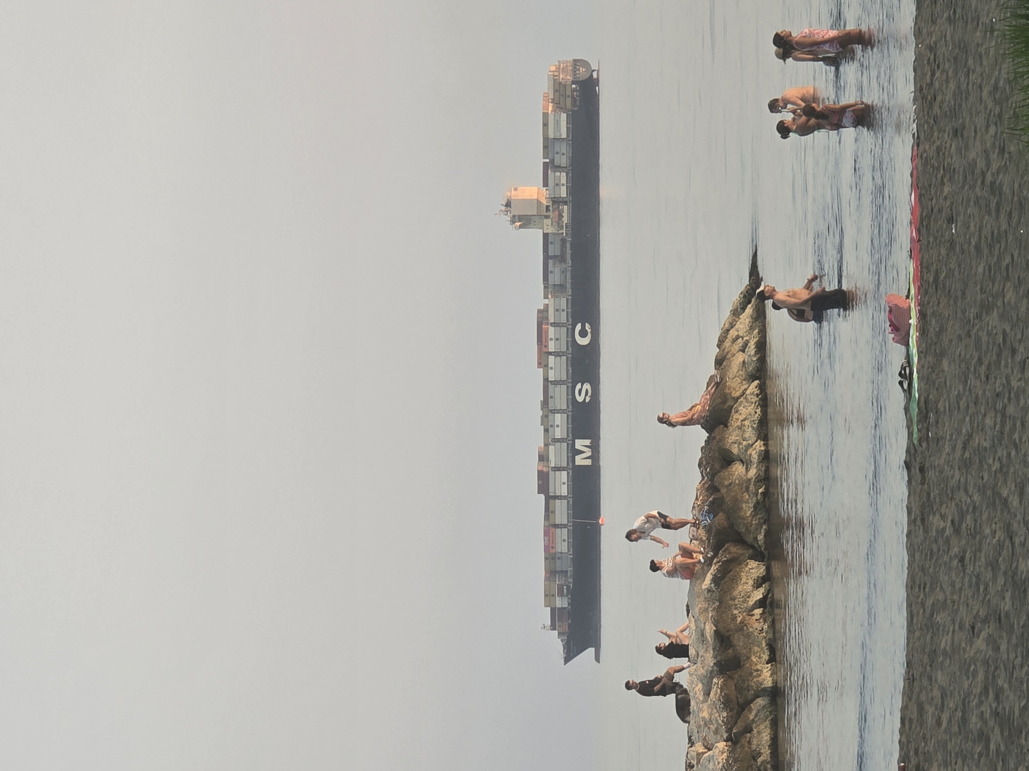 An outdoor, eye-level, medium shot captures a serene coastal scene with a cargo ship in the distance. The sky is a hazy, pale gray, and the sea is a calm, muted blue. In the foreground, a sandy beach is visible, with a patch of green grass on the far right.
A rocky outcrop juts out into the water, serving as a resting spot for several individuals. On the rocks, a group of five people are scattered, with some sitting and others standing. A man in a dark t-shirt and shorts is seated with a dog beside him. To his right, a woman in a black top and shorts looks at her phone. Further along, two individuals, one in a patterned shirt and shorts, the other in a white t-shirt and dark shorts, are engaged in conversation. To the far right, a woman in a patterned dress is seated.
In the water, closer to the beach, a group of seven people are wading. Some appear to be talking or interacting, while others are simply enjoying the water. A man in a baseball cap is visible in the water. Further out, a large cargo ship, identifiable by the "MSC" logo on its side, sails across the horizon. The ship is laden with numerous shipping containers of various colors. The overall atmosphere is one of leisure and calm. The scene suggests a location along the coastline of Málaga, Spain, likely a public beach area.