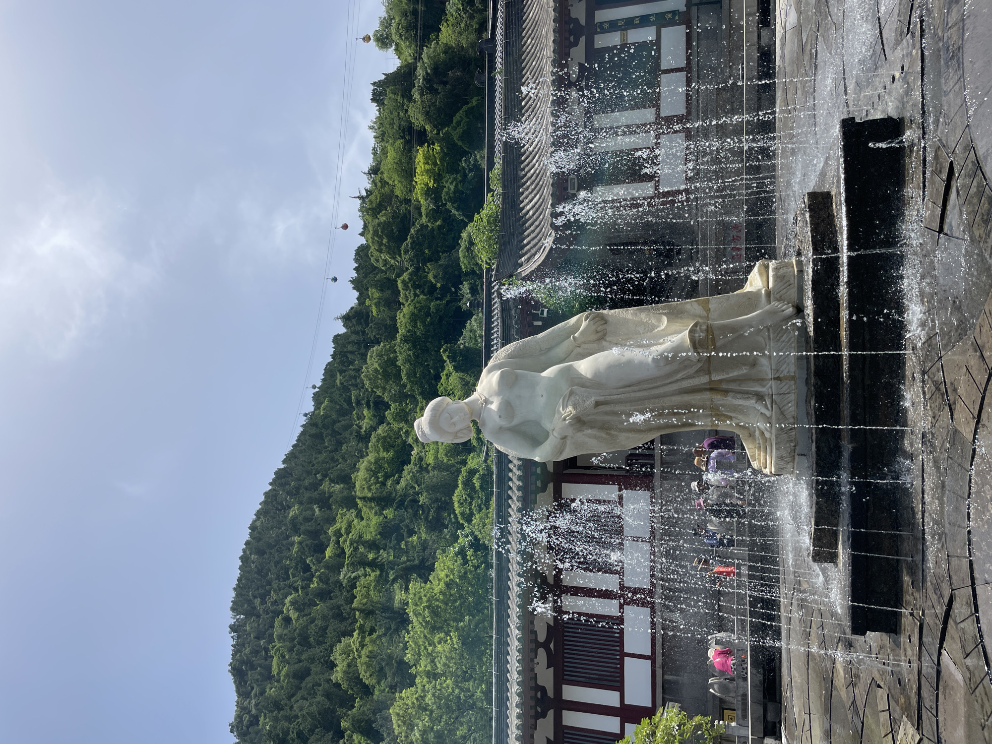 The image shows a white marble statue in a fountain in Lintong District, China. The statue depicts a woman in draped clothing. Water sprays from the fountain around the statue. In the background, there is a forested hillside and buildings with traditional Chinese architecture. A cable car line runs up the hillside. People can be seen in the background, near the base of the statue. The sky is partly cloudy.