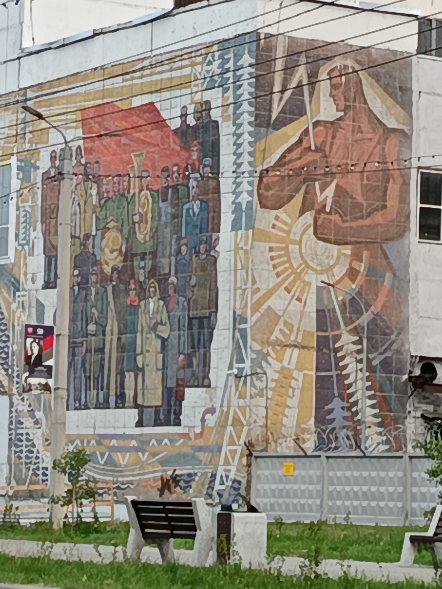 This is a full-shot, eye-level, outdoor photograph of a large, colorful mural on the side of a building in Komsomolsk-on-Amur, Russia. The mural depicts a collection of figures and symbolic imagery, rendered in a stylized, somewhat geometric manner.
On the left side of the mural, a group of people are gathered, some holding flags and musical instruments, including a tuba. They are dressed in clothing that suggests a formal occasion or a gathering of workers. A red flag is prominently displayed at the top left of this section. To the right of this group, a large, muscular figure is depicted in profile, holding what appears to be a spear or a long staff. This figure is set against a backdrop of abstract sun-like and geometric patterns. The colors in the mural range from earthy browns and reds to blues, greens, and yellows.
In the foreground, a green grassy area is visible with a concrete bench and a small tree. A street lamp is positioned above and to the left of the mural. A fence with barbed wire runs along the bottom of the mural, separating it from the foreground. A yellow warning sign is attached to the fence. The building itself is light-colored, with visible windows. The overall impression is a monumental work of art, likely commemorating a historical event or celebrating the spirit of the city or its people.