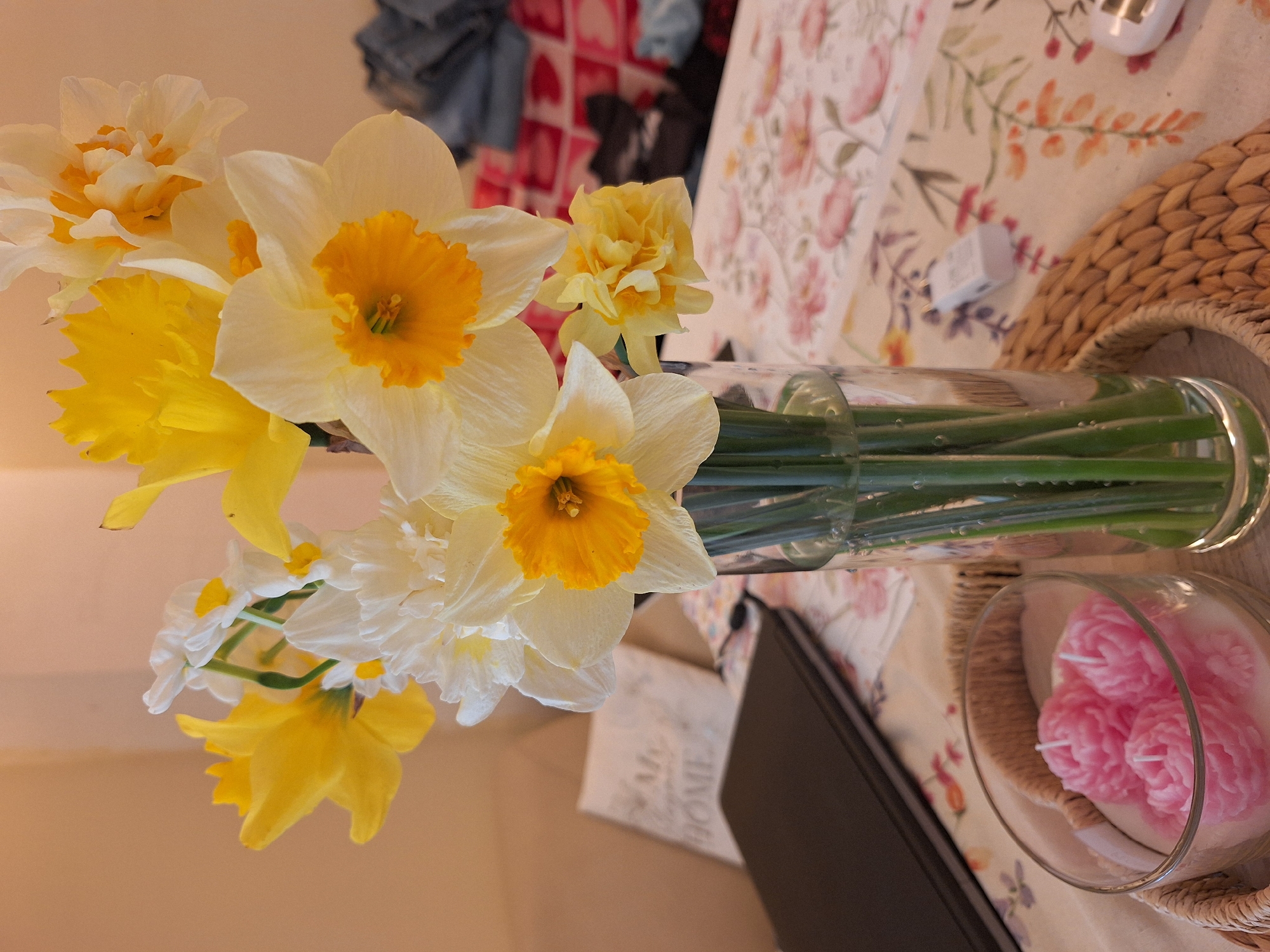 The image depicts a still life scene. A glass vase filled with water holds an assortment of daffodils in varying shades of yellow and white. The flowers are in full bloom and appear fresh. The vase sits on a tablecloth with a floral pattern. Next to the vase, there's a round wicker tray containing a glass bowl. Inside the bowl are pink floral-shaped candles. A black object, possibly a book or a notebook, is also visible. The background is blurry. The image was likely taken indoors. There is no information available to establish any connection to Varna, Bulgaria.
