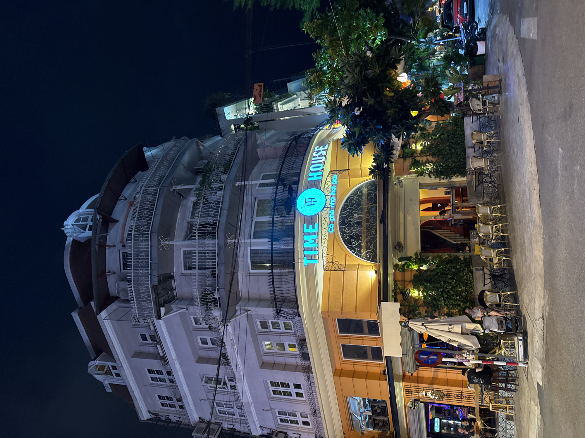 At night, a multi-story building with a rounded corner facade is illuminated. The building has several floors with balconies and windows. The ground floor is painted orange and houses a cafe with outdoor seating. The cafe has tables, chairs, and umbrellas set up on the sidewalk. Several people are seated at the tables. A neon sign above the cafe reads "TIME HOUSE" in blue lettering. The street in front of the cafe is paved, and a red car is parked on the right side of the image. The scene appears to be on a street in Nha Trang, Vietnam, as indicated by the cafe's name, "TIME HOUSE COPHEN NHA THOI GIAN", which translates to "TIME HOUSE COFFEE TIME".