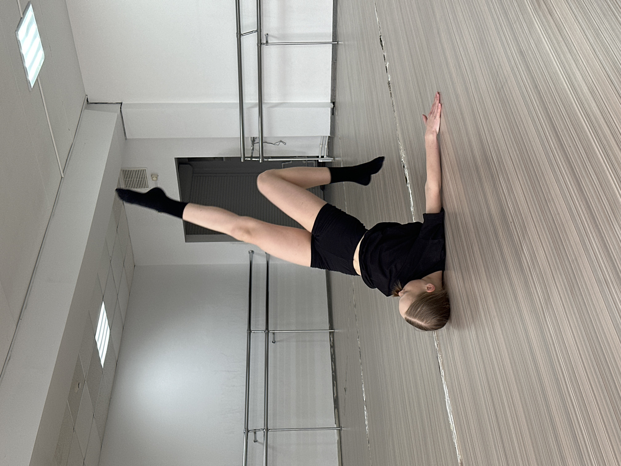 A young female dancer in black clothing (shorts, top, socks) is positioned horizontally in a dance studio or gymnasium, possibly in Votkinsk, Russia, though the exact location cannot be confirmed from the image alone. She is leaning against a light gray, vertically striped wall, with her left arm extended upward and her right leg bent at the knee and crossed over the left leg. The left leg is extended straight and upward. Her head rests against the wall. The floor is made of the same light gray, vertically striped material. In the background are silver ballet barres attached to the wall and a closed dark gray or black door. The room has a white ceiling with recessed lighting.