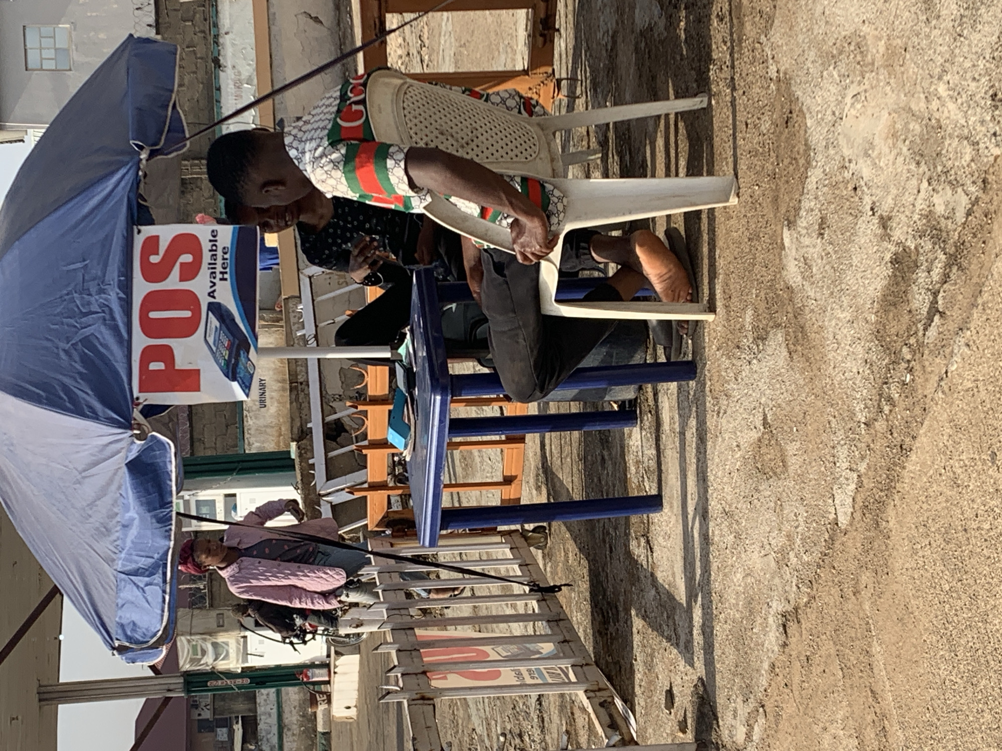The image shows an outdoor scene in Rafin Par, Nigeria, featuring two men sitting under a blue and white umbrella displaying a "POS Available Here" sign. One man is sitting on a white plastic chair and the other is sitting on a blue plastic chair. A woman in a pink jacket is walking in the foreground. The ground is a mixture of concrete and dirt. The setting appears to be a semi-urban commercial area with surrounding buildings.