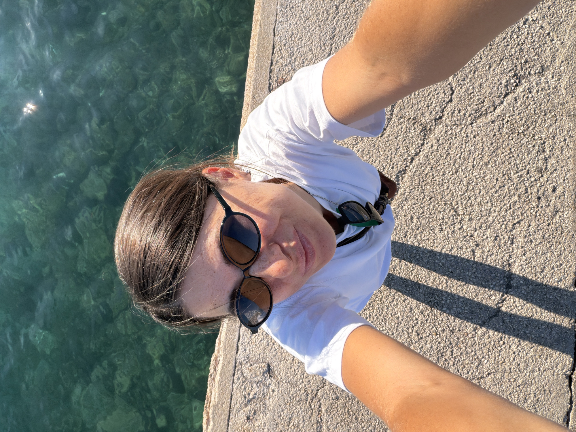 A high-angle, over-the-shoulder selfie shows a woman wearing a white t-shirt and sunglasses, standing on a concrete promenade next to clear, turquoise water. The water appears to be the Adriatic Sea, with visible rocks and sunlight reflecting on the surface. The woman is looking up towards the camera, with a slight smile. Her dark hair is pulled back, and she has freckles on her nose and cheeks. A necklace with a pendant is visible around her neck, and a pair of sunglasses is clipped to her shirt. The promenade has a rough, textured surface with visible cracks and two long shadows cast by an unseen object, suggesting it is a sunny day. The scene is likely a popular waterfront area in Budva, Montenegro, given the characteristic clear water and promenade style.