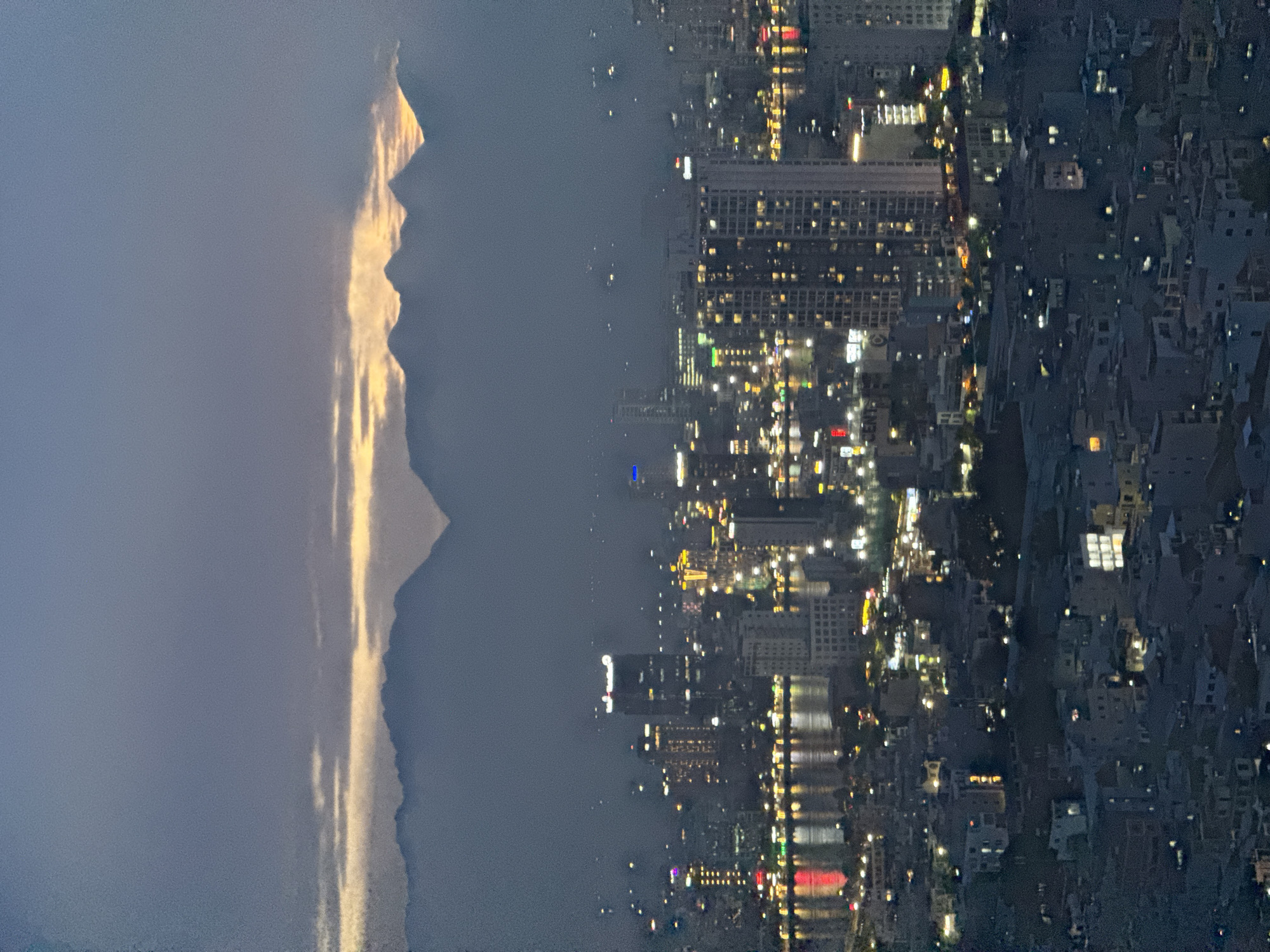 The image is an aerial, nighttime view of a city with a mountainous silhouette in the background. The city is illuminated with countless lights from buildings and streetlights, suggesting a densely populated urban area. The lights reflect on a body of water that runs through the city, creating shimmering streaks of color. The mountains are dark and imposing, with a sliver of light on their peaks, possibly from the setting or rising sun. The sky is a deep blue, transitioning to black at the top of the image.
The scene appears to capture the transition from day to night, or vice versa, with the warm glow on the mountains contrasting with the cool artificial lights of the city. There is no visible activity of people, but the illuminated buildings imply daily life. The overall mood is serene and perhaps a little awe-inspiring, given the juxtaposition of nature and urban development.
The image is oriented vertically, with the mountains on the left and the city on the right. The reflections on the water are particularly striking, with hints of red and yellow lights visible. The crops provided highlight the sharp, jagged outline of the mountains and the intricate network of lights in the city. Some text is visible on building signs, such as "CENTRAL" and what appears to be "FB" in the city view, but it is mostly illegible due to the distance and resolution. Based on the provided location context, this scene is likely Đà Nẵng, Vietnam, where the city is known for its coastal setting and proximity to mountains.