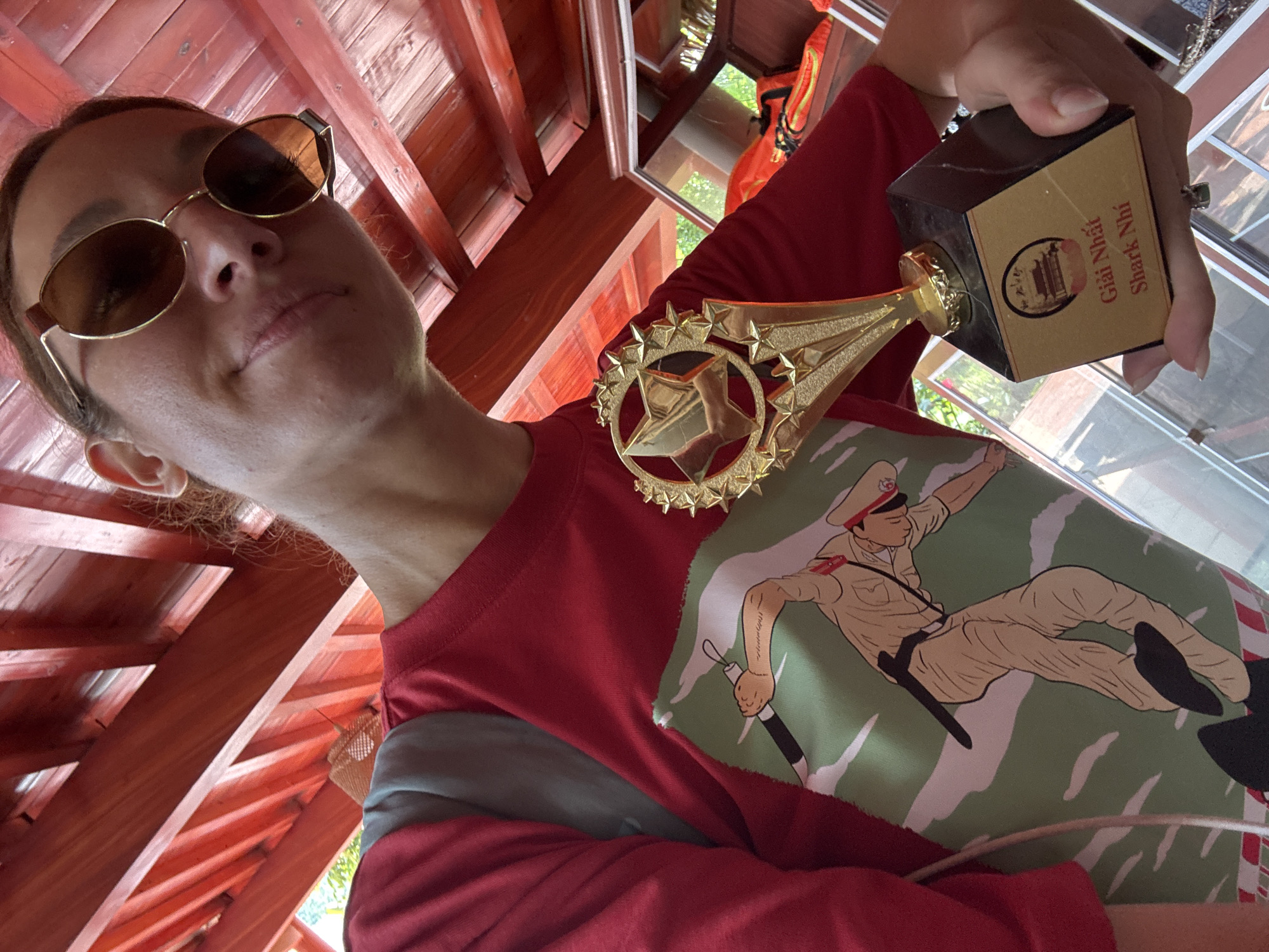 A person, likely a woman, is captured in a low-angle selfie, looking slightly upwards with a subtle, satisfied expression, wearing reflective sunglasses and a red long-sleeved shirt. Her shirt features a distinctive graphic print depicting a cartoon-style figure in a khaki military-like uniform, complete with a cap and red insignia, set against a green and white camouflage background. In her right hand, she proudly holds up a golden trophy, which has a star emblem at the top and a black and gold base.
The trophy's base displays Vietnamese text: "Giải Nhất Shark Nhi" (First Prize Shark Nhi), along with a small illustration of a traditional Vietnamese building (possibly a pagoda or temple). The setting appears to be indoors or under a sheltered area with a reddish-brown wooden plank and beam ceiling, suggesting a rustic or traditional architectural style. Reflections in the background indicate windows or openings to the outside, revealing glimpses of greenery. The natural lighting suggests the photo was taken during the daytime. The location context indicates this is in Thông Nguyên Commune, Vietnam. The person's pose and the trophy indicate an event of achievement or celebration.