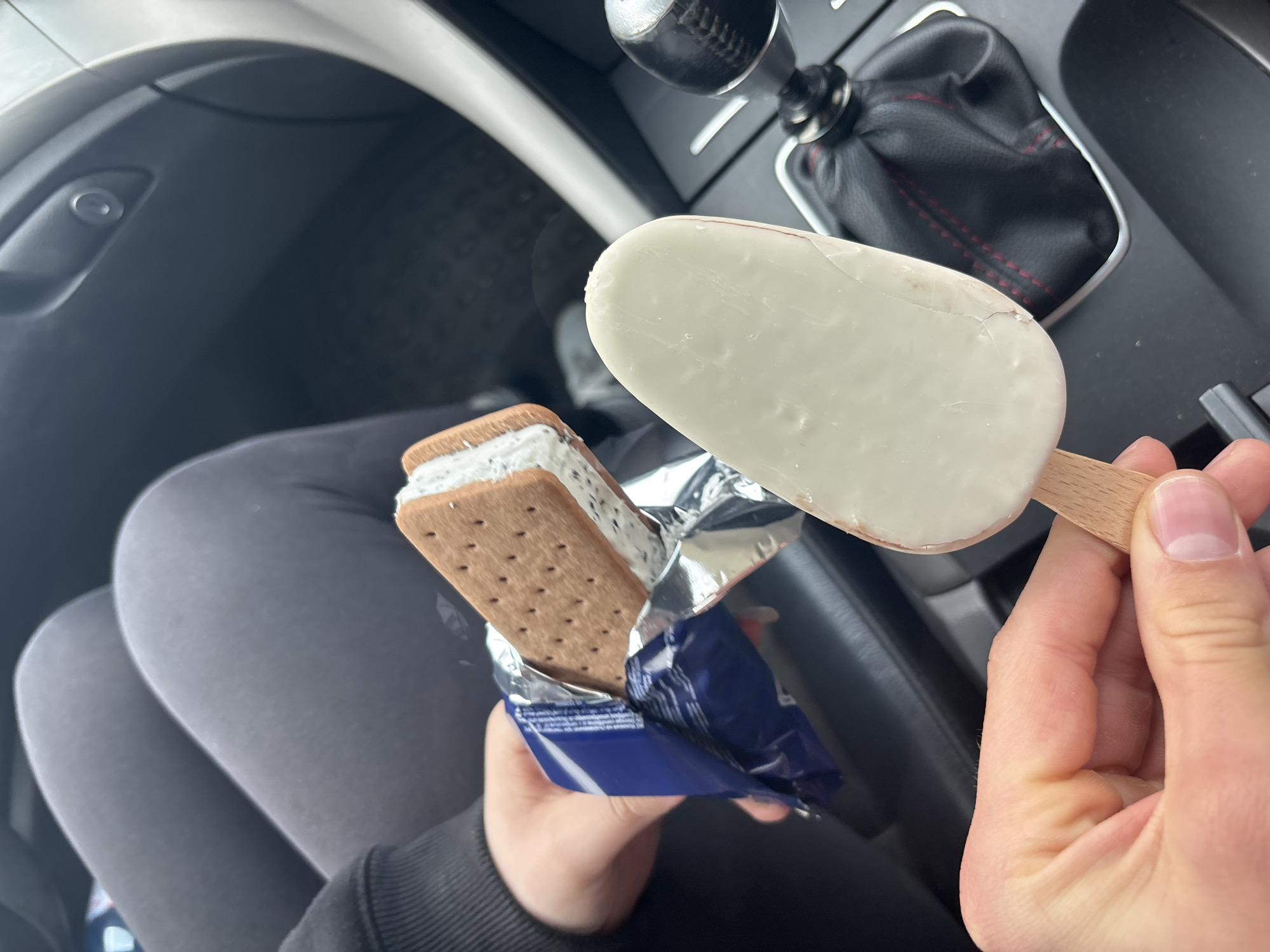 This close-up image captures a person's hands holding two different ice cream treats inside a car, likely in Dobromirtsi, Bulgaria. The primary subject is a person, whose hands are prominent in the foreground, holding the frozen desserts. Their legs, clad in dark grey leggings, are visible in the lower-left background, suggesting they are seated.
One hand holds a rectangular ice cream bar on a wooden stick, coated in smooth white chocolate, which shows a few minor cracks on its surface. The other hand holds a partially unwrapped ice cream sandwich. This sandwich features a thick layer of white ice cream, speckled with dark inclusions (possibly cookies and cream), nestled between two rectangular, perforated brown cookies. The ice cream sandwich is still partly encased in its blue and silver foil packaging, which has some unreadable text visible.
The setting is clearly the interior of a vehicle. Details of the car include a black leather gear shift boot with distinctive red stitching, indicating a manual transmission, and elements of the dashboard and door panel, including a small round button on the door. The lighting suggests it is daytime. The activity taking place is the preparation to consume these treats. No explicit emotions are visible, but the context implies enjoyment of a snack.