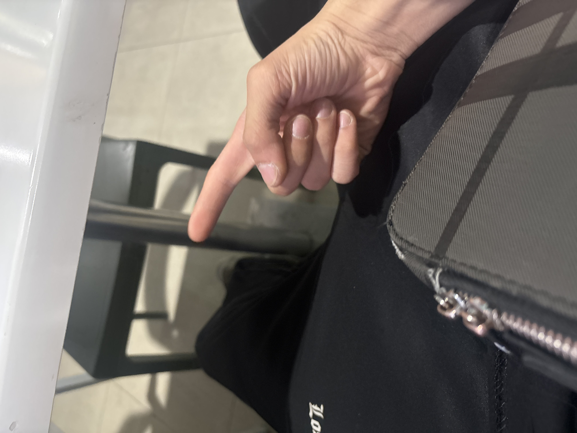 The image is a close-up, angled shot from an overhead perspective, capturing a person's hand and surrounding elements in an indoor setting in Yerevan, Armenia.
The primary subject is a person's right hand, seen from above, with the index finger extended downwards in a pointing gesture. The hand shows visible skin texture and natural, unpolished fingernails. A portion of the person's leg or lap, dressed in dark, possibly athletic-style pants or leggings, occupies the lower and right parts of the frame. Resting on the lap is a grey, textured bag or wallet featuring a prominent silver zipper and what appears to be a smaller, metallic zipper pull.
The background elements suggest an indoor public space, such as a cafe or waiting area. To the left, a bright white, vertical structure, possibly a table leg or partition, is visible. Behind the pointing hand, a dark metal frame, likely part of a chair or table base, can be seen, casting subtle shadows on the light-colored, possibly tiled floor.
The pointing gesture indicates an action of drawing attention to something or providing direction. The lighting is bright and even, typical of an indoor environment during the day or with good artificial illumination. On the dark garment on the person's lap, a partially visible white text, which appears to be a rotated brand name such as "LOTTE" or "KOTTE," is discernible.