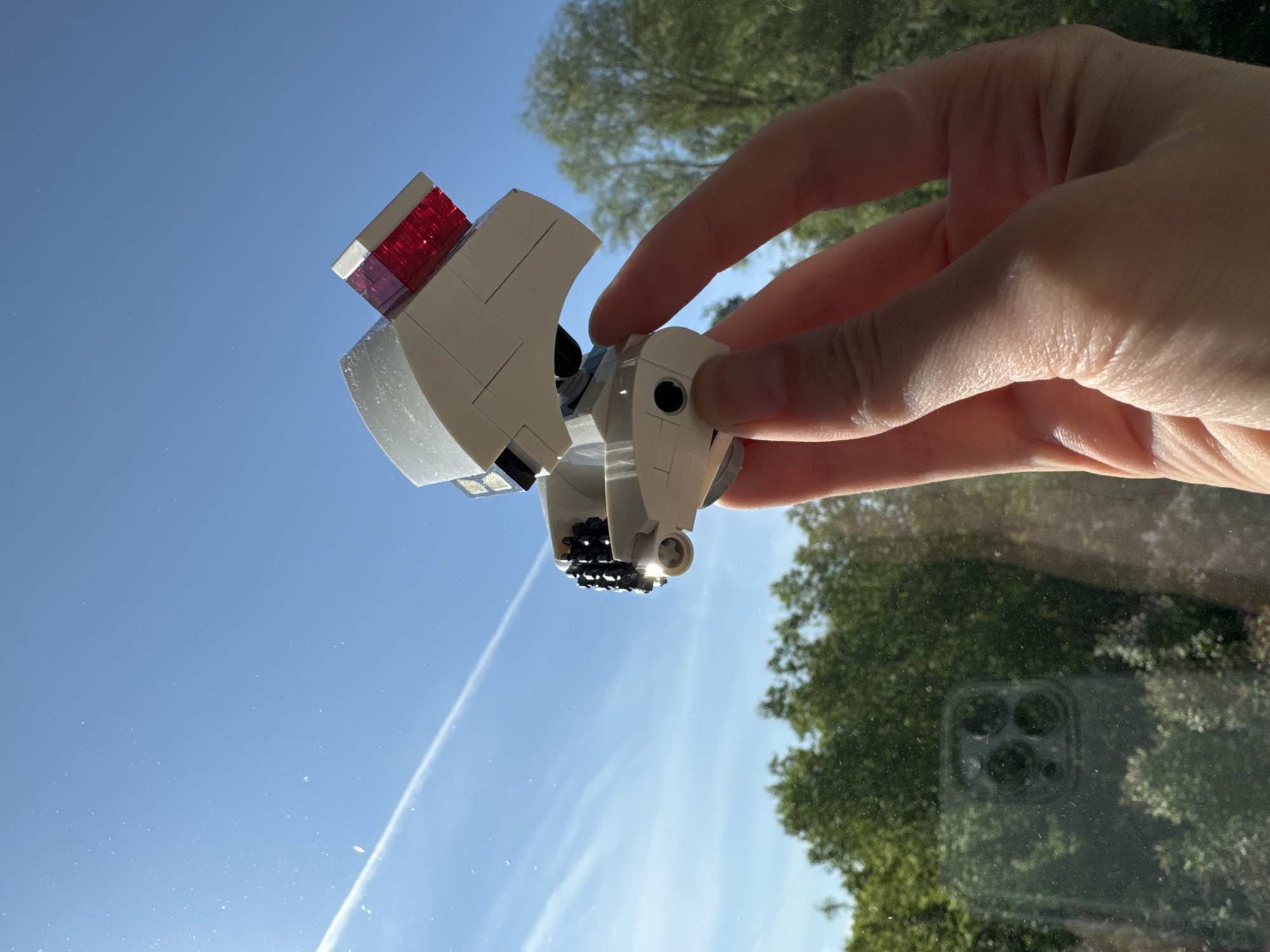 The image, taken in Riga, Latvia, features a human hand holding a small, custom-built white LEGO model against a bright blue sky. The LEGO construction appears to be a stylized vehicle or spaceship, with red transparent bricks visible on one end and black studded details on its underside. The hand holds the model at an upward angle, silhouetted against the clear sky, which shows a distinct white contrail from an aircraft.
In the background, out-of-focus green trees are visible, suggesting an outdoor setting or a view through a window. The presence of numerous small dust specks and a clear reflection of a smartphone's triple camera array on the lower right side of the image indicates that the photograph is taken through a glass surface, likely a window. The time of day appears to be bright daylight, with strong natural light illuminating the scene. No discernible text or specific emotions are visible.