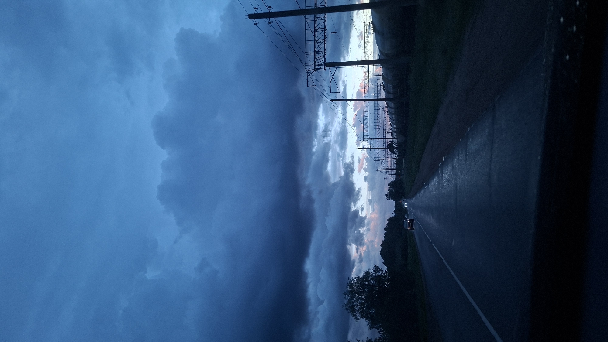 A photograph taken in the city of Смалявічы, Belarus, shows a long, wet asphalt road with a white line marking its center. A single vehicle with lit headlights is visible on the road. To the right of the road is a green field. Above the field are power lines supported by metal poles. Above the road and field, the sky is dramatically divided between a dark blue stormy cloud formation on the left and a lighter sky with hints of pink and gray on the right.