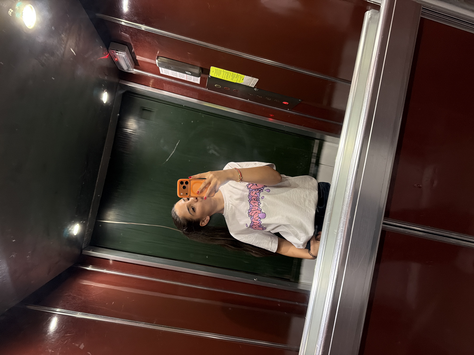 The image is an interior shot of an elevator with a woman taking a selfie in the mirror. The elevator walls are dark red. The elevator doors are a polished silver. The elevator interior features a panel with buttons and a light fixture. The woman is wearing a white t-shirt with a graphic print and has long, dark hair pulled back in a ponytail. She is holding an orange smartphone to take the picture. The precise location in Sofia, Bulgaria is not discernible from the image.