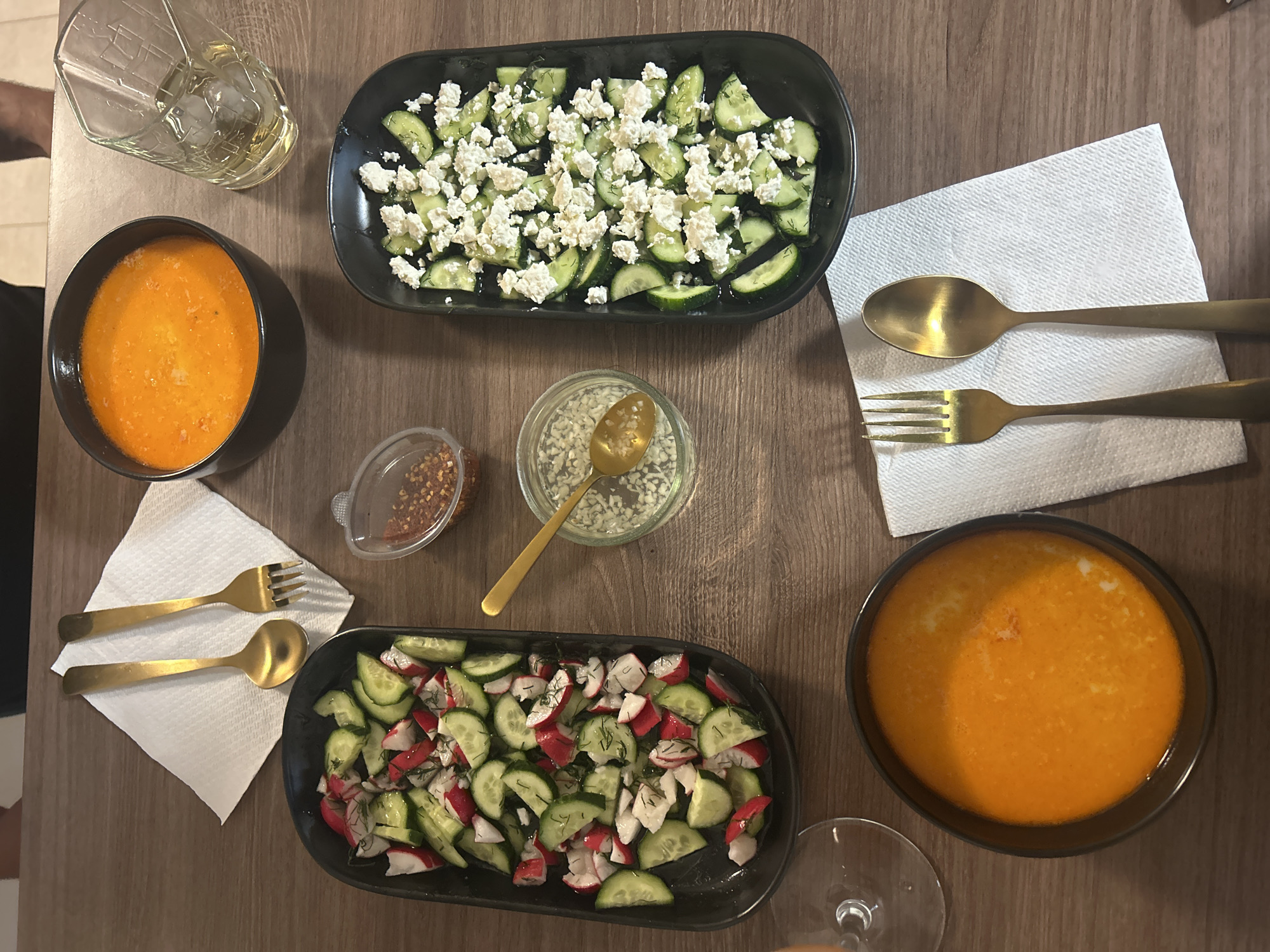 The image depicts a top-down view of a wooden table with a meal set up for two people. Two bowls of orange-colored soup are visible on opposite sides of the frame. One is accompanied by a clear glass with an unknown liquid and ice. The other is near the base of a wine glass. Two black oblong serving dishes contain salads. One salad consists of sliced cucumbers topped with crumbled white cheese, possibly feta. The other salad is a mix of sliced cucumbers and radishes, garnished with dill. In the center, there's a small container of red pepper flakes and a small jar filled with a white dressing and a golden spoon. Two sets of golden-colored utensils, each consisting of a fork and a spoon, are placed on white napkins. There is no indication that the location is Veliko Tarnovo, Bulgaria. There are no people visible in the image, only their implied presence through the prepared meal. The environment is indoors, suggested by the table setting.