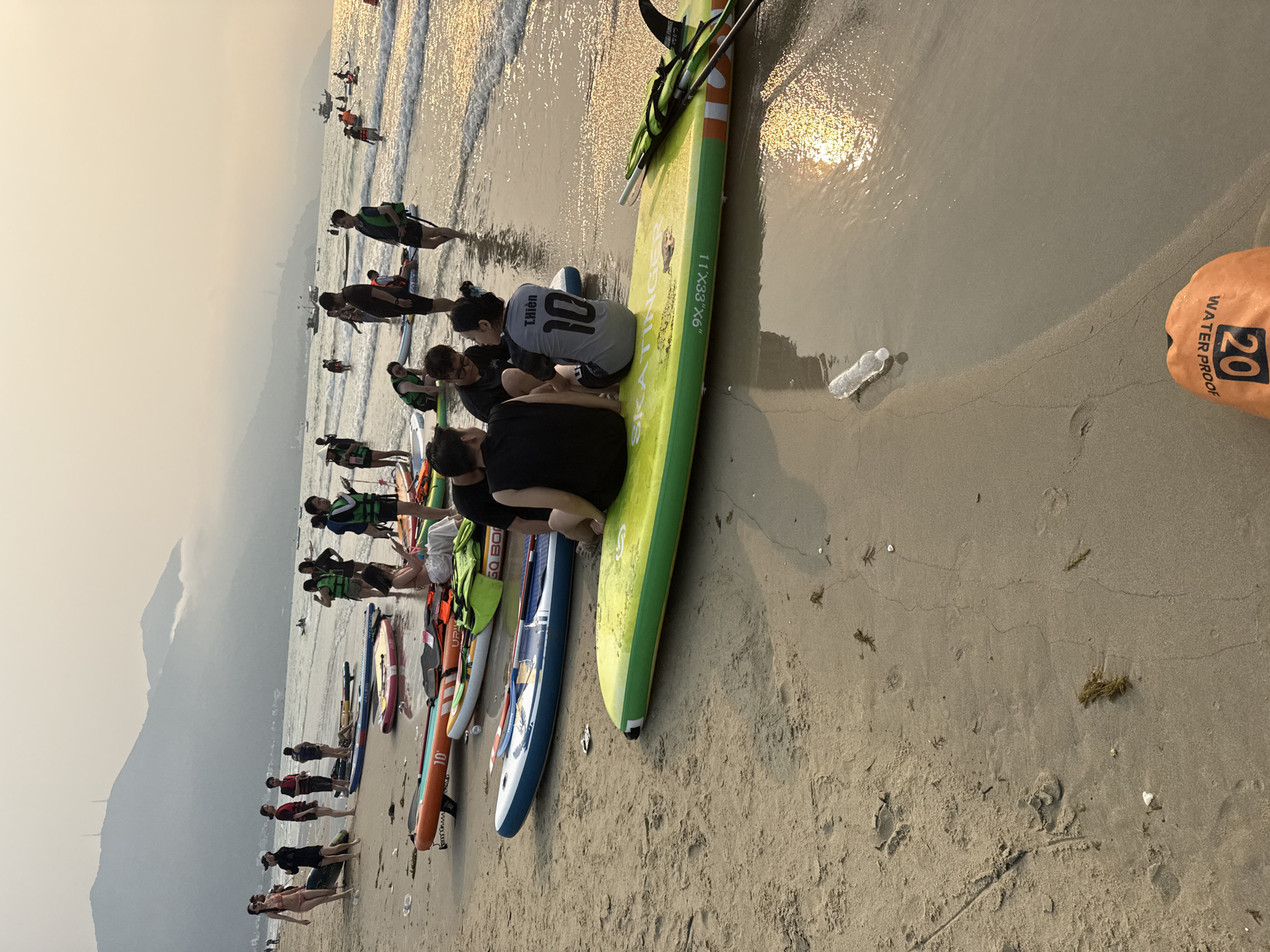 The image depicts a beach scene, likely at sunset given the hazy lighting conditions and low sun angle. The location is the coastal city of Đà Nẵng, Vietnam, indicated by the mountain visible in the background resembling the Đà Nẵng mountains. A group of people are either preparing for or have just finished stand-up paddleboarding. Several paddleboards of various colors and sizes are lying on the beach. Some individuals are standing or walking with life vests on, while others are sitting on the paddleboards. One person has a shirt with the name "T.Hiền" and the number "10" on the back. In the foreground, a green paddleboard with dimensions "11'X33"X6"" is visible. A plastic water bottle and a waterproof bag with the number "20" are on the sand. The sea is calm with small waves breaking on the shore. The sand is wet and slightly reflective.