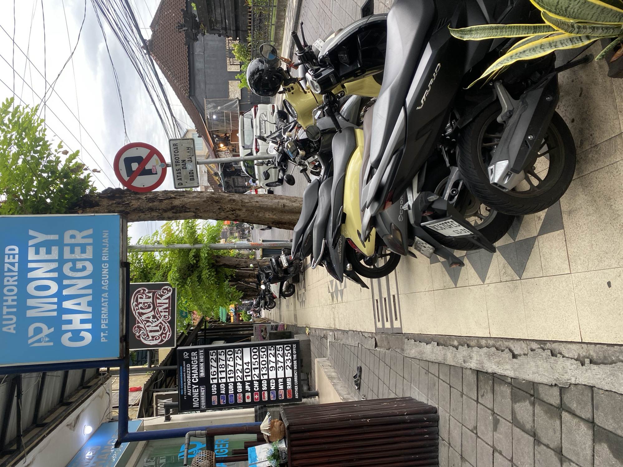 The media file captures a street scene in Kuta, Indonesia, characterized by parked motorcycles lining a sidewalk next to a road under an overcast sky during daytime. The image is taken from a slightly elevated perspective, looking down the street.
The primary subjects are numerous scooters, predominantly black and yellow, parked closely together on a tiled sidewalk. A "No Parking" sign, a red circle with a diagonal line over a black 'P', is prominent on a pole along with a rectangular sign displaying text that reads "D ATAS TROTOAR DAN BADAN JALAN," likely indicating parking regulations on the sidewalk and road. Overhead, a dense network of electrical wires crisscrosses the sky.
On the left side of the frame, several signs for a money changer are visible. A large blue sign announces "AUTHORIZED MONEY CHANGER PT. PERMATA AGUNG RINJANI." Below it, a black sign with white text lists exchange rates for various currencies: USD 16725, AUD 11875, EURO 19750, GBP 22725, JPY 104, CHF 21500, SGD 13050, NZD 9725, MYR 3725, and RMB 2350, with a note "ANOTHER CURRENCY INSIDE THE OFFICE." Another smaller sign indicates "NO COMMISSION." Nearby, a decorative sign for "GRACE BANK" is also seen.
A small wooden table or bench is positioned near the money changer signs, and a single sandal lies on the sidewalk. Further down the street, more parked vehicles, including a white car, are visible. Green foliage from trees and plants adds to the streetscape, including a snake plant in the foreground.