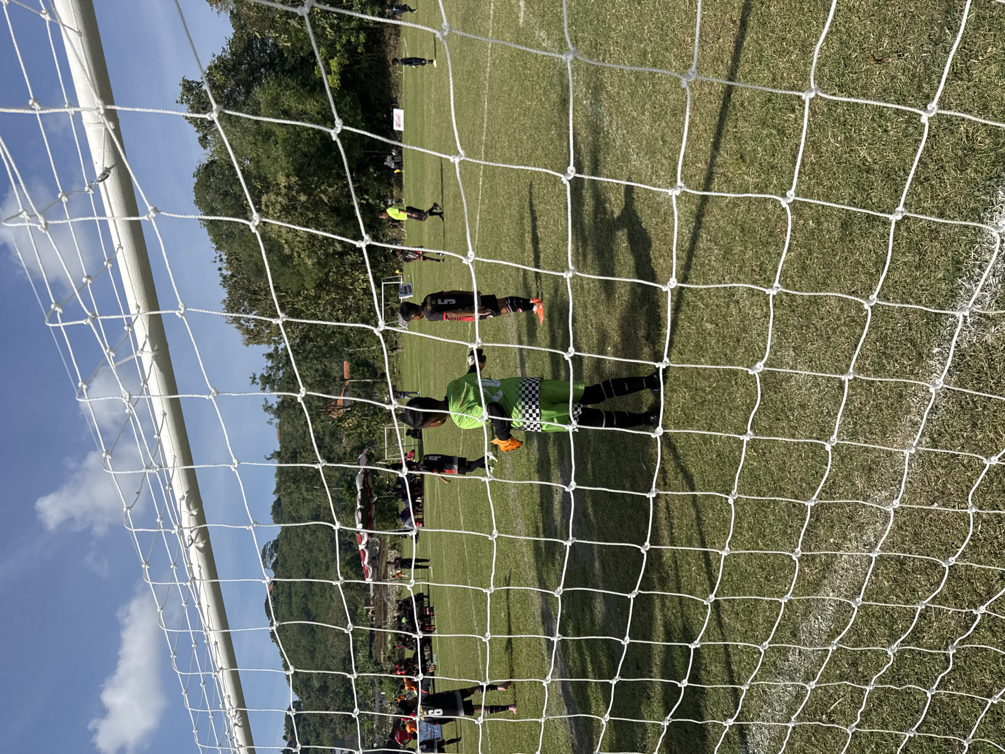 A low-angle shot, taken from inside a soccer goal, looks out onto a soccer field in Kuta Selatan, Indonesia. The white net of the goal is in the foreground, partially obscuring the field. A goalkeeper, dressed in lime green with black and white checkered sleeves, is positioned in front of the goal. Other soccer players in black and orange uniforms are scattered across the field. A crowd of people is visible in the background along with some trees and structures. The sky is blue with scattered clouds. The image is rotated, so the scene appears sideways.