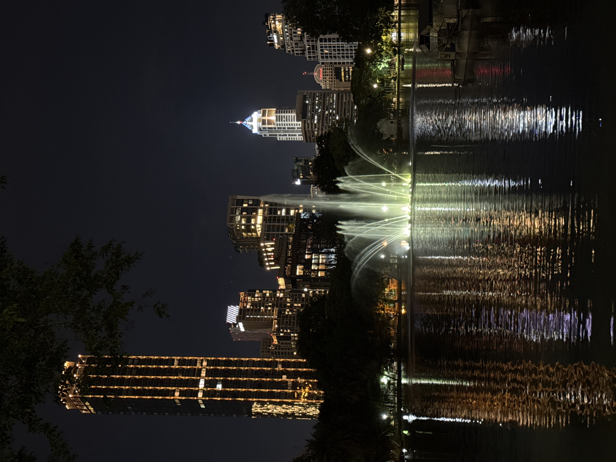 Here is a concise description of the image:
The image shows a nighttime view of the Bangkok skyline reflected in a body of water, likely a lake or pond, with a fountain in the foreground. The skyscrapers are illuminated with lights. The water reflects these lights, creating a shimmering effect. The location is likely Lumphini Park in Bangkok, Thailand, known for its water features and views of the surrounding skyline. There are no people visible. The focus is on the urban environment, the interplay of light, and the water.