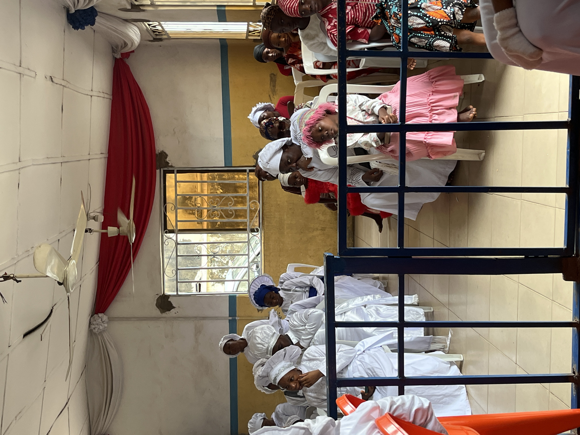 The image depicts an indoor gathering, possibly a religious service, with approximately 30 individuals, predominantly women and children, seated in rows of white plastic chairs. The majority of attendees are wearing white attire, including headdresses, with some children also in colorful dresses and headscarves. A young girl in a pink dress and a pink wig sits in the foreground. The room has a high, white-tiled ceiling with two ceiling fans. Red and white drapery is hung along the top of the walls, and a large window with metal bars is visible in the background. The floor is tiled in a neutral color. A blue metal fence or barrier runs across the middle ground of the image. The overall impression is of a formal assembly within a simple interior space.