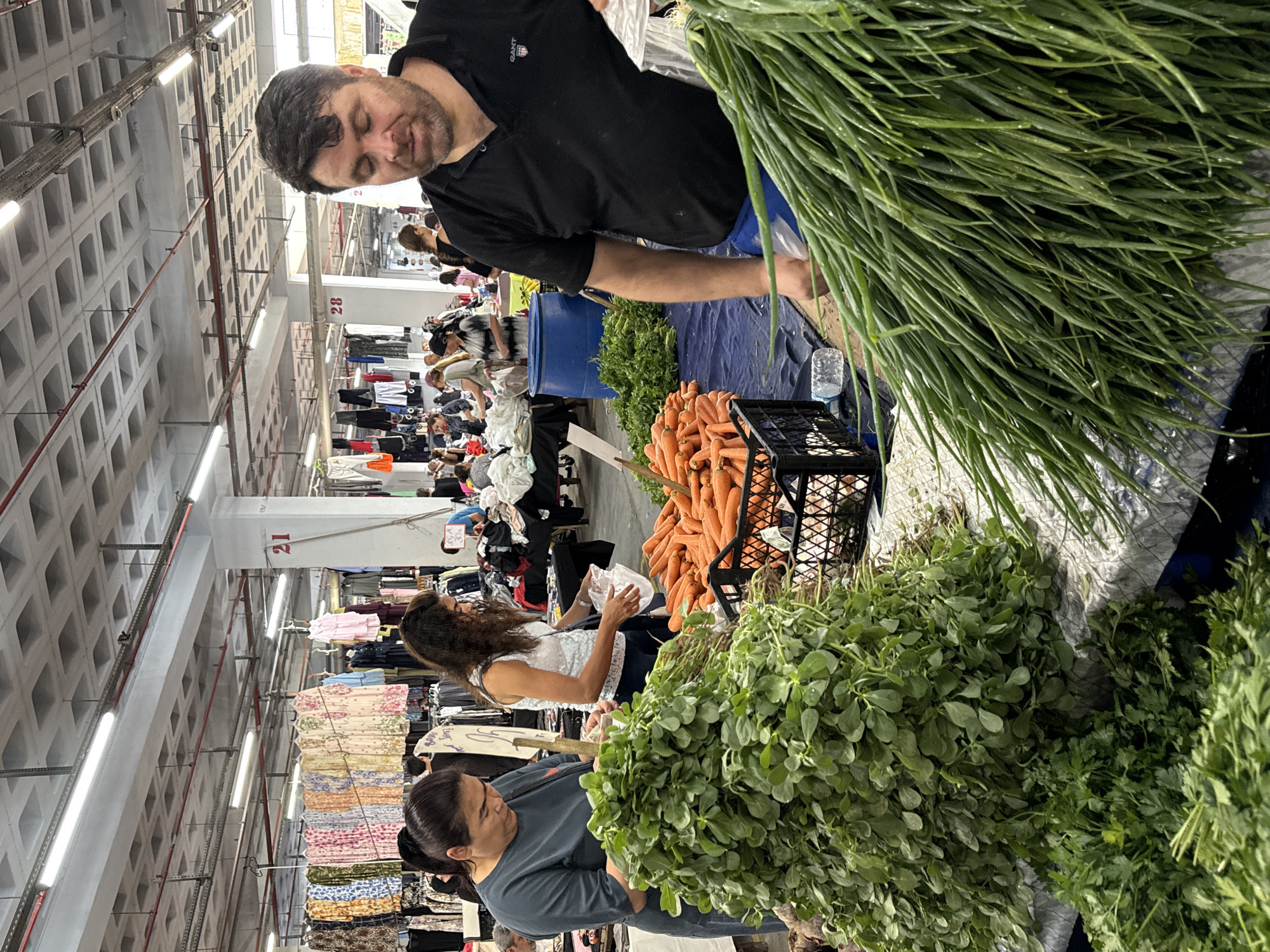 The image depicts an indoor market scene in Kadıköy, Turkey, likely a produce market. In the foreground, a man wearing a black polo shirt and blue shorts is standing behind a display of fresh green onions. To his left, two women are seen browsing vegetables. One woman, wearing a grey top, is examining a bunch of leafy green vegetables, possibly purslane. Another woman, in a white patterned top, is holding a plastic bag and looking towards the vegetables. A crate of bright orange carrots is visible between the women and the man. In the background, stalls with various clothing items are displayed, suggesting a multi-purpose market. The ceiling is a grid structure with fluorescent lighting. The overall atmosphere is that of a bustling marketplace.