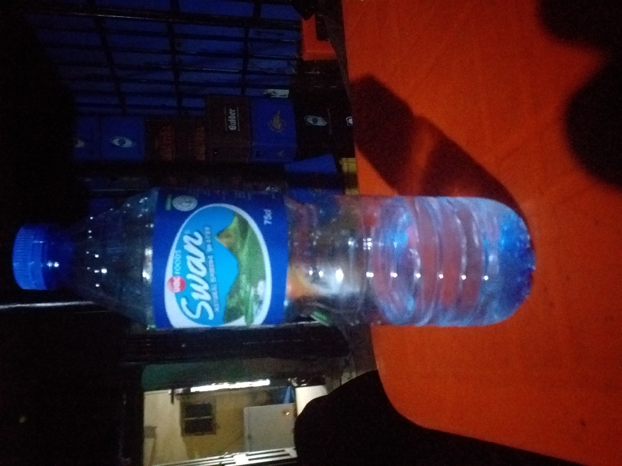 A bottle of Swan Natural Spring Water is positioned on an orange surface. The bottle, which is partially filled with water, has a blue cap and a label featuring the Swan logo and a depiction of mountains. In the background, several blue crates are stacked, and to the left, a doorway reveals a glimpse of a room with windows and a white appliance. The scene appears to be indoors or in a covered outdoor area, with shadows suggesting artificial lighting.